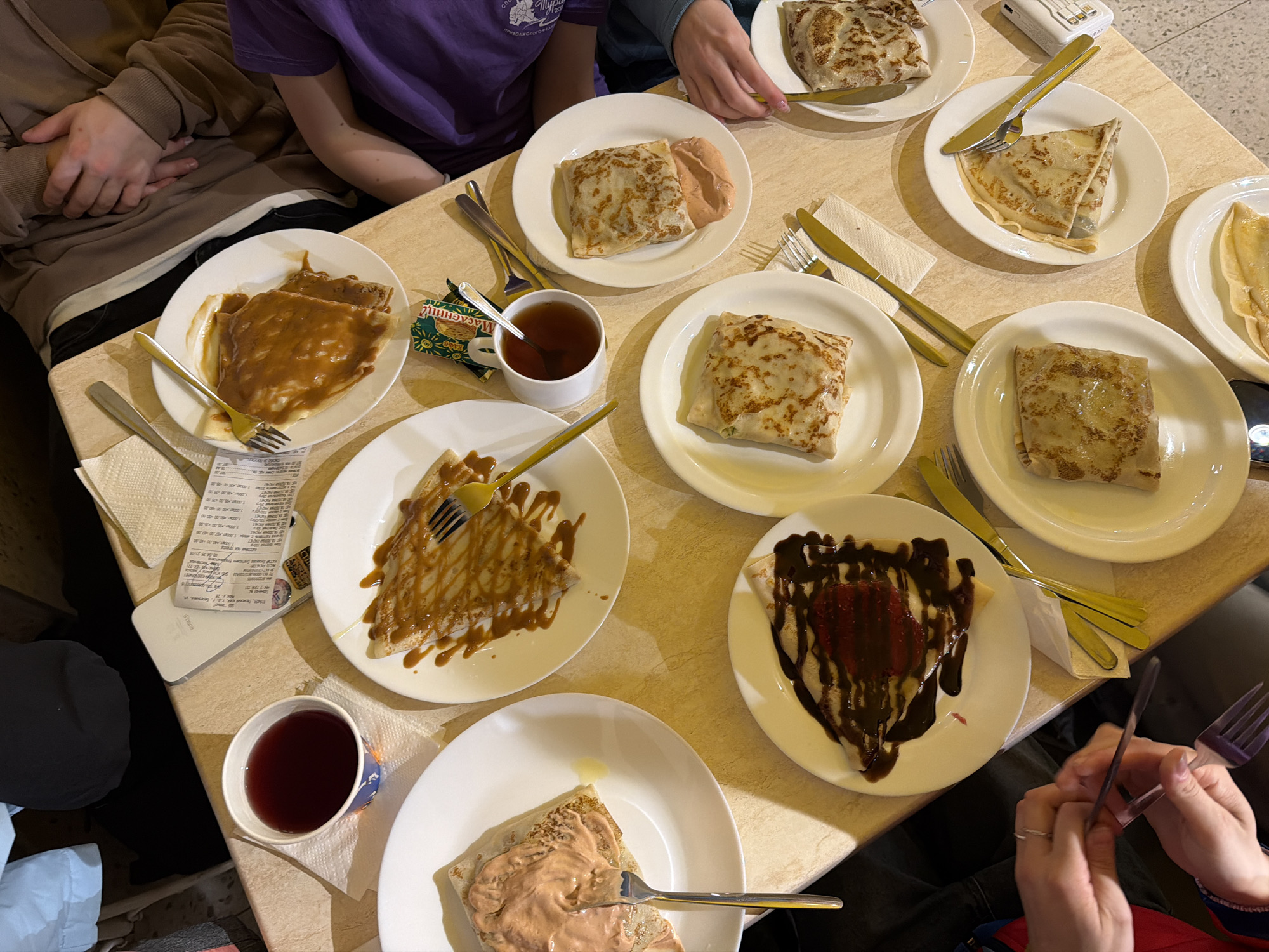 The image depicts a table in what appears to be a restaurant in Berezniki, Russia. Several people are seated around the table, and there are plates of food. The dishes consist of crepes with a variety of fillings or toppings. These toppings include a caramel-like sauce, a chocolate and berry topping, and a meat-based filling. Tea is also present in cups on the table. Receipts, cutlery, and other dining accessories are present on the table. The room lighting appears to be bright, and the overall scene conveys a casual dining experience.