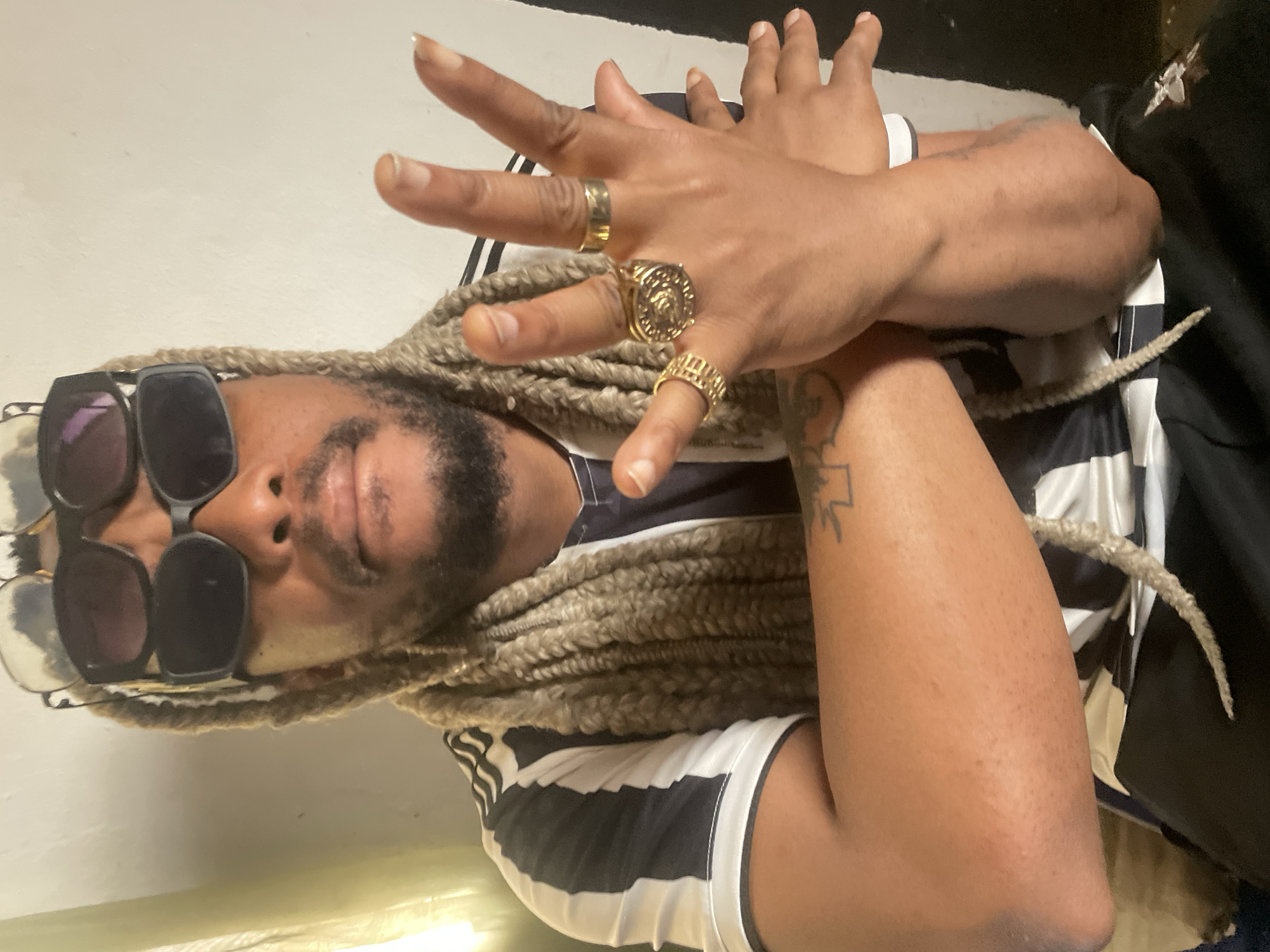 A man with blonde braided hair, sunglasses, and gold rings poses for a portrait. He is wearing a black and white striped shirt. The location in the city of Jos, Nigeria, is unknown. The background is a plain wall.