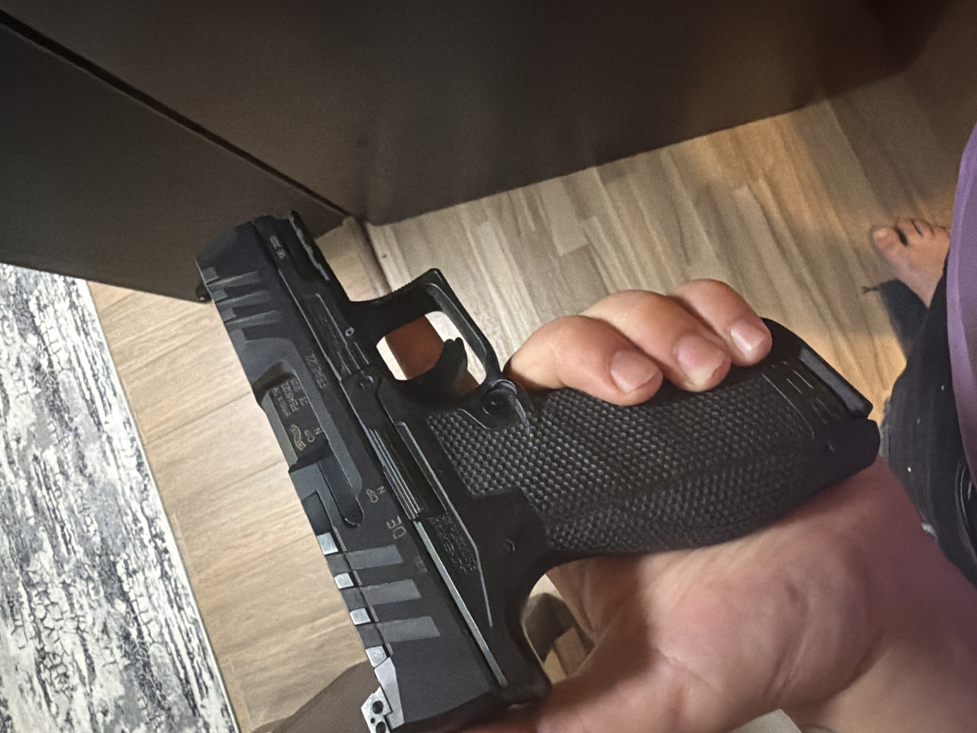 A person's right hand firmly grips a black semi-automatic handgun, holding it upright. The handgun is predominantly black with textured grips, and features visible markings on its slide. Among the discernible text are "FS4022", "9x19", and "DE", alongside a QR code-like pattern and other proof marks or serial numbers. The weapon's trigger is visible within the trigger guard.
The scene is indoors, with a dark, solid surface, possibly the underside of a desk or table, visible in the upper portion of the frame. Below the handgun, the floor is covered with light-colored, wood-patterned flooring. A portion of a grey and white patterned rug is also visible on the left side. In the background, a person's bare feet are visible, suggesting a relaxed, domestic setting. The lighting appears to be ambient indoor light.