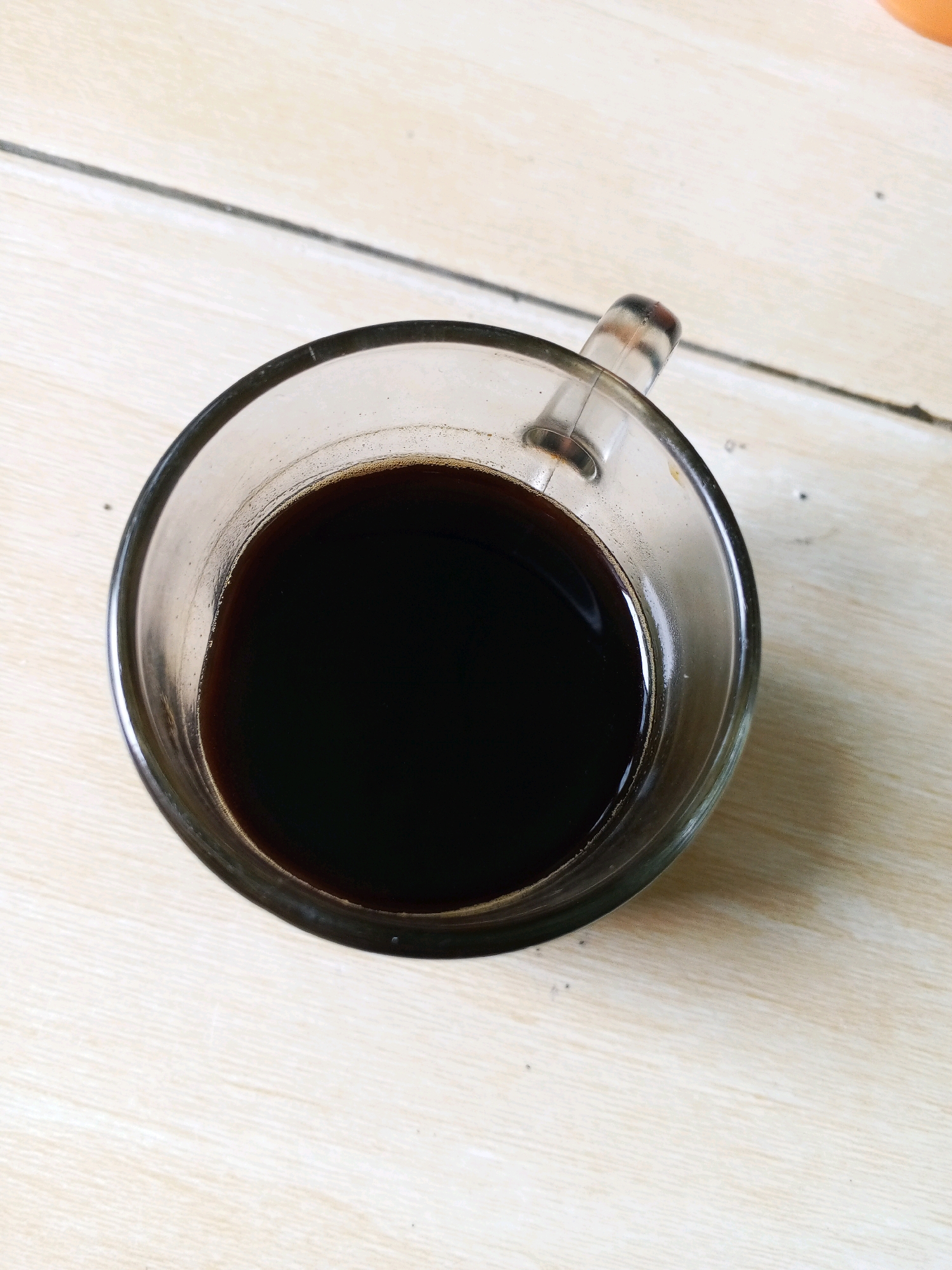 A close-up, overhead shot shows a clear glass mug filled with dark coffee, placed on a light-colored, tiled surface. The mug has a simple handle. The coffee is dark brown and appears to have a slight foam or crema on the surface. The tiled surface is made of rectangular tiles with visible grout lines. The lighting is natural, casting subtle shadows. The scene is likely indoors, possibly on a table in a home or cafe in Deli Tua Timur, Indonesia.