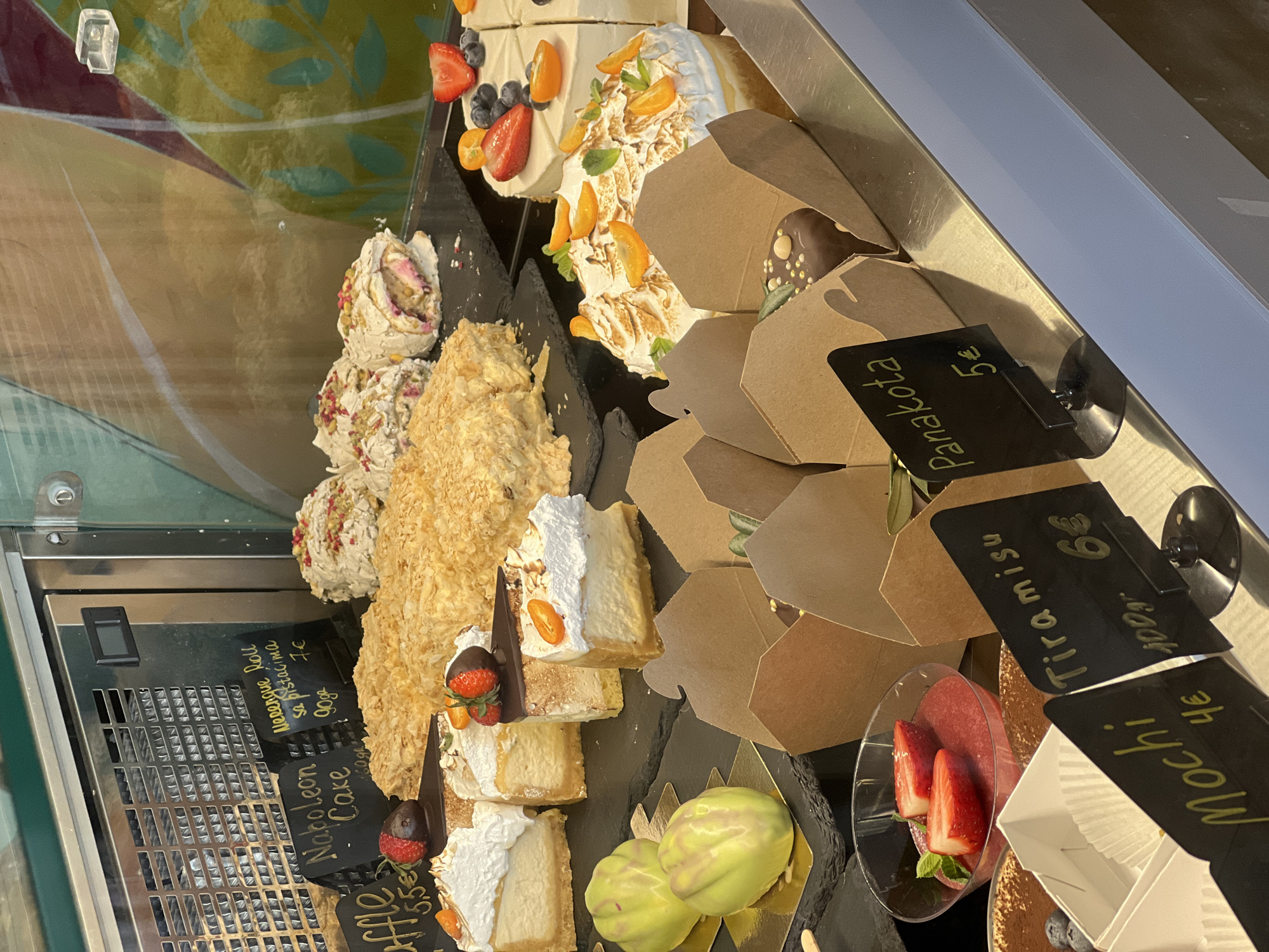 The image shows a display case in what appears to be a bakery or dessert shop, possibly located in Rafailovići, Montenegro, based on the text recognition of "sa pistacima." The case is filled with various cakes, pastries, and desserts, each labeled with a price.
There are slices of cake decorated with fruit, meringue rolls, Napoleon cake, and individual servings of desserts packaged in brown boxes and small containers. Specific items are labeled "Mochi 4€," "Tiramisu 6€," and "Panakota 5€." Other handwritten labels indicate "Affle 5,5€," "Naboleon Cake," and "Merengue Roll sa pistacima 7€." The desserts include components such as strawberries, blueberries, chocolate, and cream. The items are attractively arranged for sale.