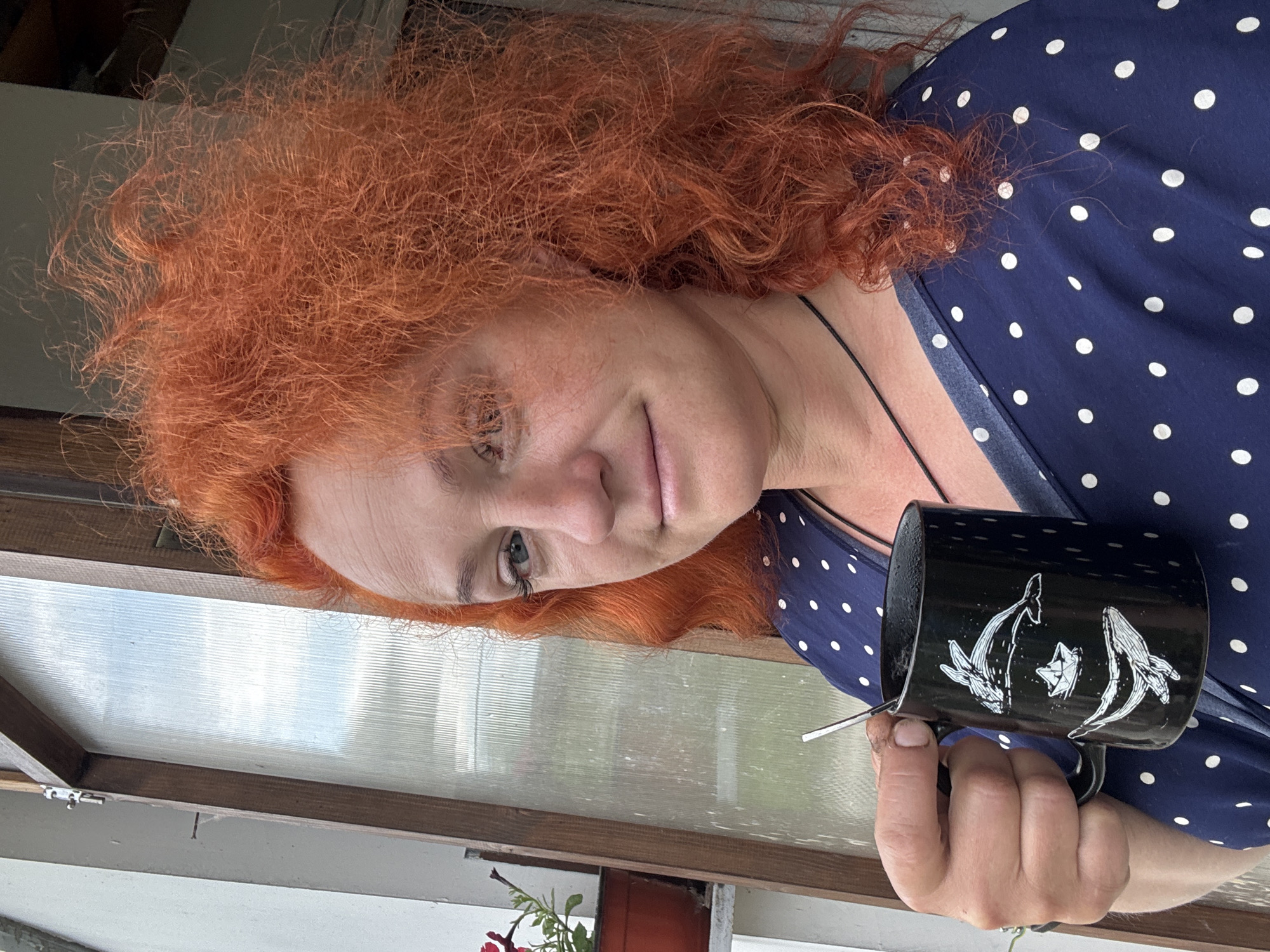 A woman with vibrant red, curly hair, wearing a dark blue dress with white polka dots, holds a black mug with a white drawing of whales and a paper boat. She is standing in front of a window with corrugated glass panes, possibly a sunroom or a greenhouse, in Chaikovsky, Russia. A potted plant with red flowers is visible to the left of the window. The scene appears to be outdoors, with natural light illuminating the woman and her surroundings.