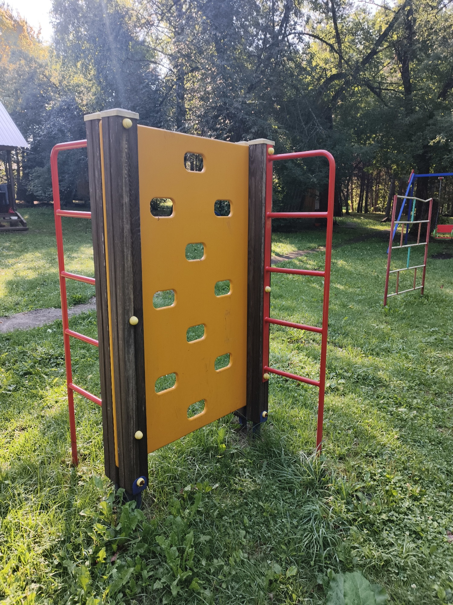 The image depicts a playground scene on a sunny day. In the foreground, a yellow climbing wall with rectangular cutouts is supported by dark wooden posts and framed by red metal bars. Yellow balls are visible on the climbing wall and its supports. To the right of the climbing wall, a partially visible ladder-like structure with red supports and green and blue rungs stands in the grass. The background features a grassy area with trees and sunlight filtering through the leaves. To the left, a portion of a white-roofed structure, possibly a gazebo or shelter, is visible. The ground is covered in green grass and some weeds. The overall setting suggests a recreational area in an outdoor environment. This scene is likely located in or near the city of Катунь, Russia, given the context. There are no people present in the image.