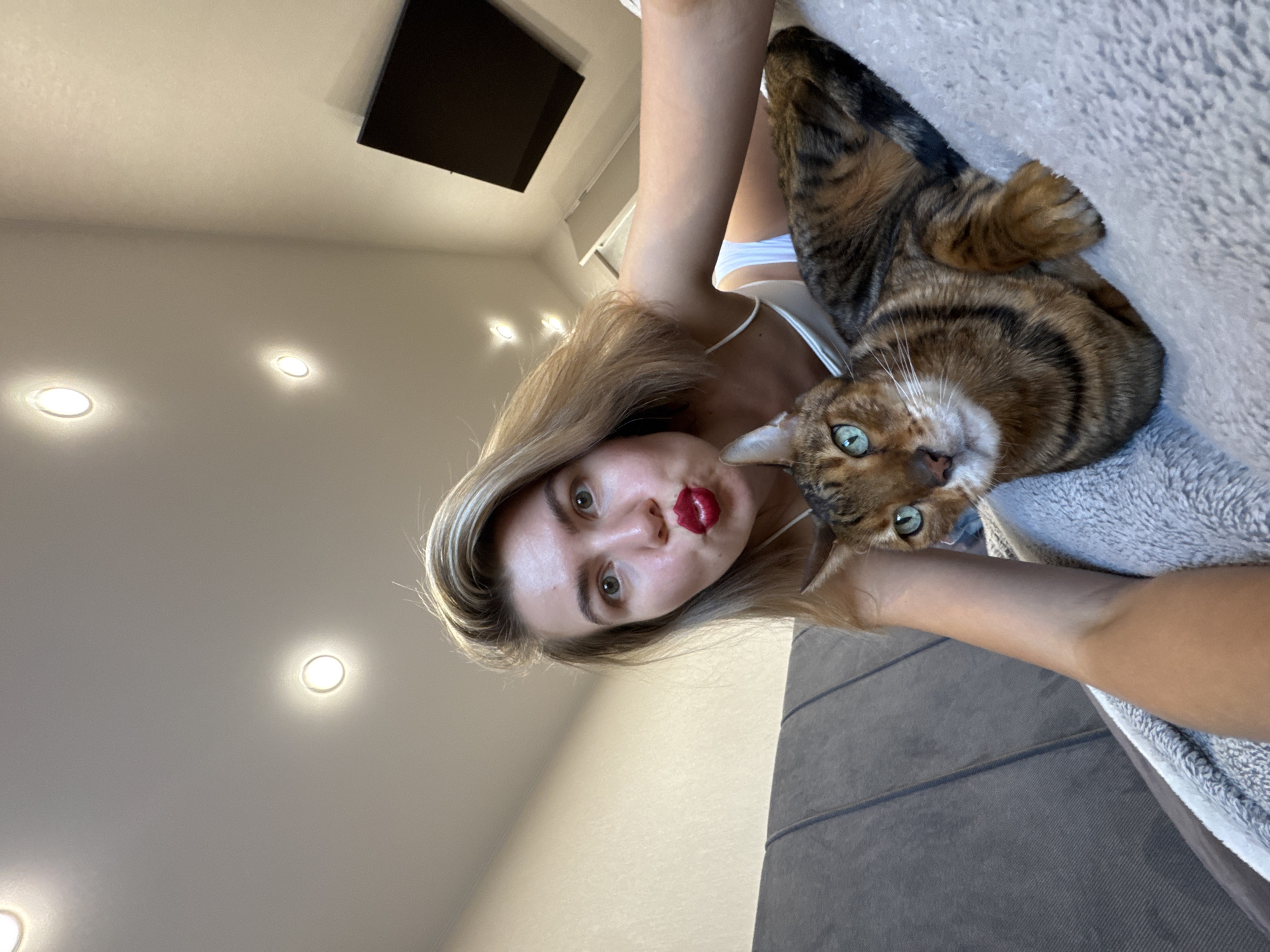 A woman with blonde hair, wearing a white top and red lipstick, is holding a Bengal cat. The location appears to be inside a residence in the city of Unknown, Unknown, as evidenced by the ceiling with recessed lighting and a black rectangular object mounted on the wall. The woman is holding the camera above her, creating an angled selfie. The cat is looking directly at the camera with green eyes.