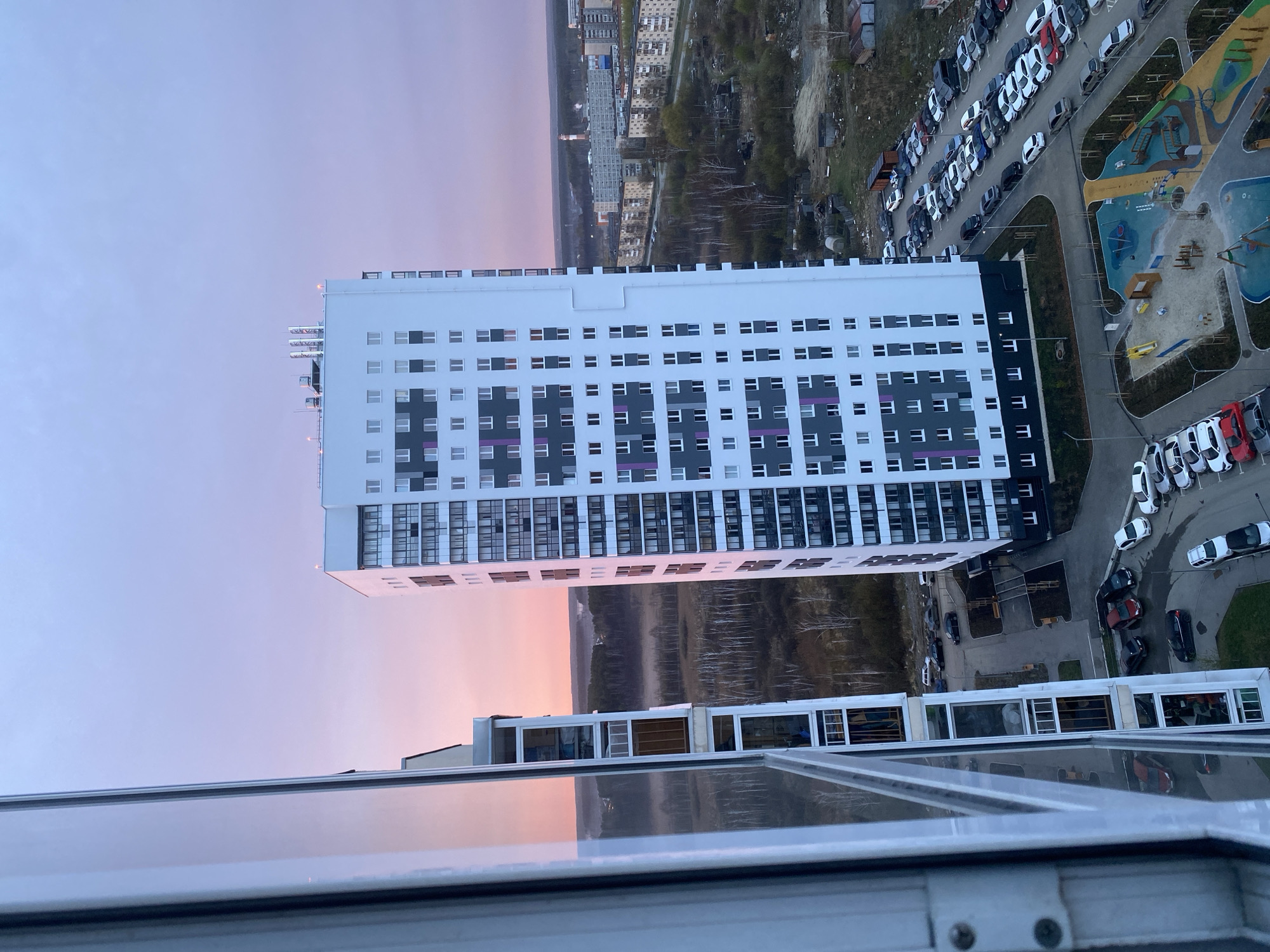 The image is an eye-level, outdoor shot taken from a high vantage point, possibly a building's window.
The main subject is a tall, modern apartment building with a white and dark gray facade. It has a regular pattern of windows, some with purple accents. The building is surrounded by other buildings, trees, and a parking lot full of cars. A colorful playground can be seen adjacent to the building.
In the distance, there is a view of the city of Yekaterinburg, Russia, with more buildings and greenery. The sky is a mix of light blue, white, and pink, suggesting either a sunrise or sunset.
The foreground shows a portion of the window from which the photo was taken, with reflections of the sky and surrounding buildings.