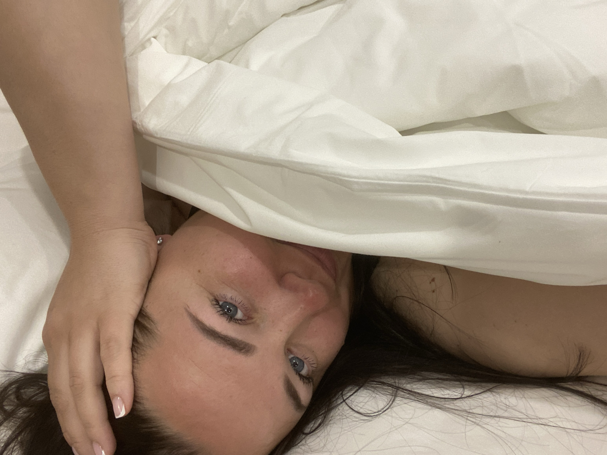 A woman with blue eyes and dark hair lies on a white bed, partially covered by a white duvet. Her left hand rests on her forehead. The setting seems to be indoors, likely a bedroom. There is no specific information in the image to identify the location as Sochi, Russia.