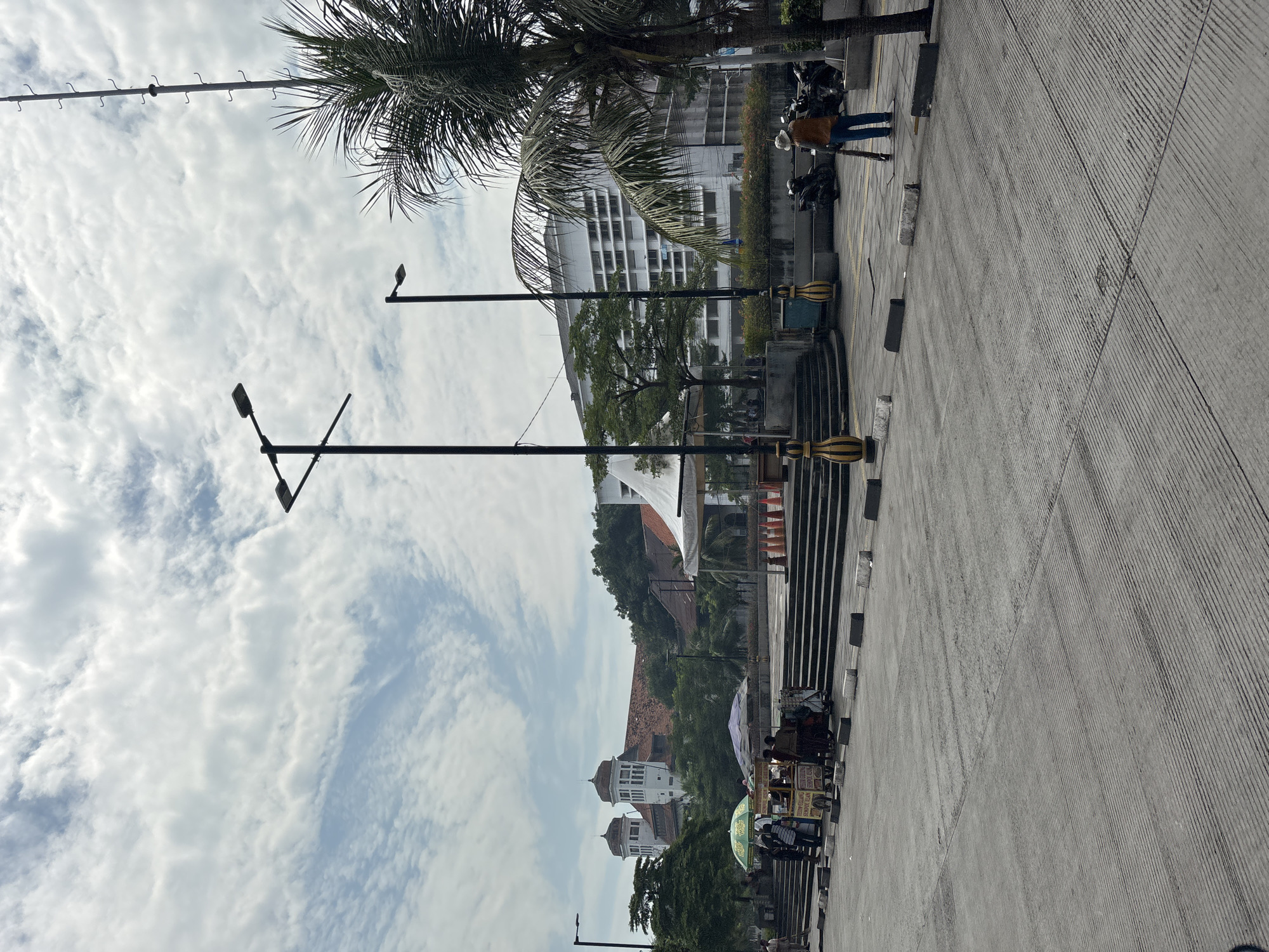 This image captures a bustling daytime scene in what appears to be an urban or semi-urban environment, consistent with West Jakarta, Indonesia. The perspective is from a slightly elevated position looking down a wide paved area, possibly a street or pedestrian zone, towards a distant hill.
In the foreground, a gray concrete or asphalt surface with linear patterns dominates, suggesting a road or large pathway. Along its right edge (after rotation), several black and yellow striped bollards or barriers are visible. Two prominent black street lamps with multiple fixtures stand vertically in the left and center of the frame, extending upwards.
Further back on the right, a person wearing a light-colored hat, brown jacket, and blue jeans walks away from the camera. Nearby, several motorcycles or scooters are parked. Adjacent to this area is a modern-looking multi-story building with numerous windows, partially obscured by lush green trees, including tall palm trees. A long metal pole with hooks, possibly for decorative lights or banners, stretches across the sky in the upper left.
Towards the midground, several mobile vendor carts or kiosks, some with colorful umbrellas, are set up along the paved area. Visible text on one cart reads "BAKSO BAKAR," indicating grilled meatballs, while another appears to offer "COKLAT KOPI" (Chocolate Coffee) and "ES TEH" (Iced Tea). A few other people are intermittently visible around these stalls.
The background features a prominent green hill. On or near the hill, older buildings with distinct architectural styles, including white structures topped with turrets or domes, stand out. A large white canopy or tent-like structure is also visible among the trees.
The sky is partly cloudy, with scattered white clouds against a lighter blue, suggesting a bright but possibly overcast day without direct harsh sunlight. The overall atmosphere appears calm and typical of daily urban life, with pedestrian activity and commerce.