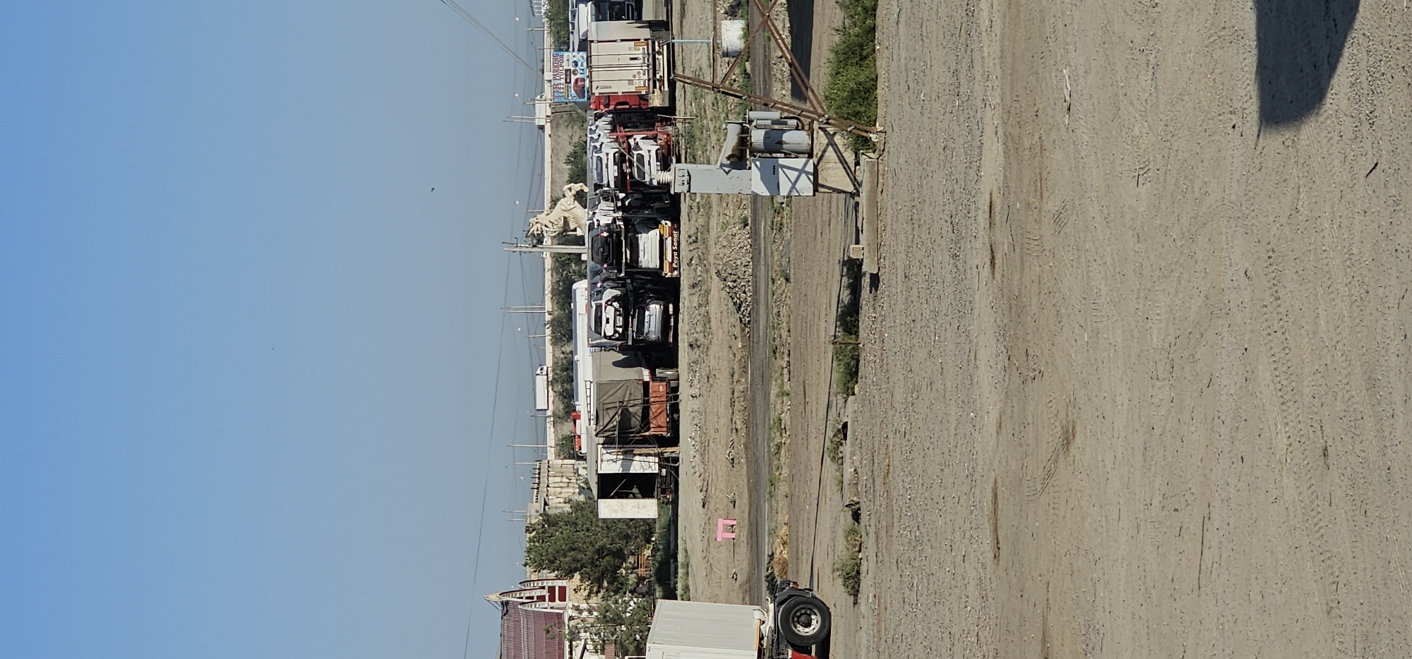 In the city of Sheyhancha, Uzbekistan, under a clear blue sky, a sunlit outdoor scene unfolds in a lot filled with vehicles and industrial equipment. The foreground is a dusty, uneven ground with tire tracks visible. To the left, a white container truck is partially visible, with its rear wheel and part of its chassis in frame. Beyond this, a small, bright pink stool rests on the dirt.
The middle ground is populated by several car carriers stacked with vehicles, and other trucks. Prominently featured is a large, light-colored statue of a horse or bull-like creature, possibly a monument, positioned behind some of the trucks. To the right of the statue, a sign with colorful graphics and text, including the words "PARKING" and "TULPON" is affixed to a pole. A large electrical transformer sits on a concrete base near the right edge of this area, connected to a metal structure that resembles a power pylon or scaffold.
In the background, a long, low-lying building with a distinctive red-tiled, arched roof is visible on the left. Further back, a series of overpasses and utility poles crisscross the horizon, with a single white truck traveling on one of the elevated roadways. Trees and bushes are scattered throughout the scene, with some appearing to be in a state of disrepair. The overall impression is of an industrial or logistical hub, possibly a storage or transit area for vehicles.