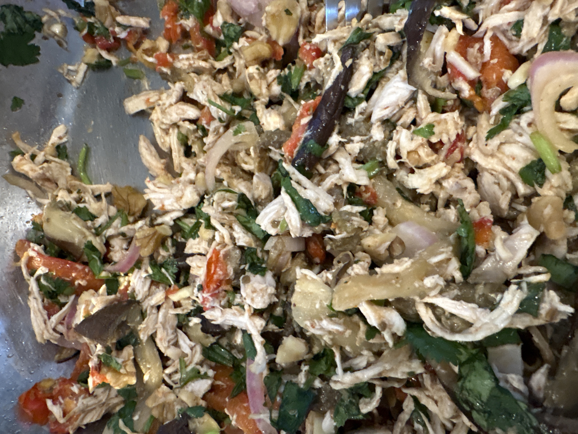 A close-up, overhead shot shows a metal bowl filled with a chicken and vegetable salad. The salad contains shredded chicken, chopped red tomatoes, green herbs, and sliced red onions. Some pieces of what appear to be eggplant are also visible. The background is out of focus and appears to be a kitchen or dining area. The image was taken in Karon, Thailand.