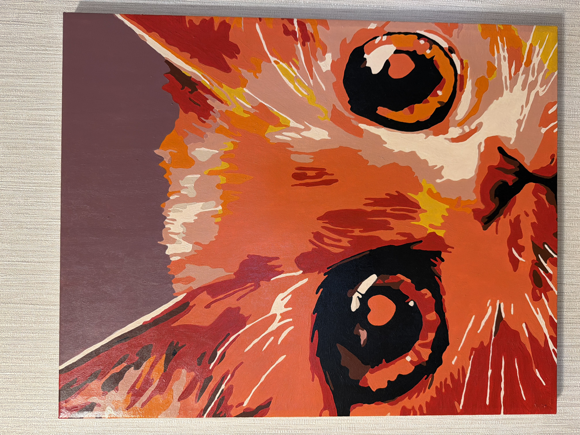 The image is an eye-level close-up of a painting of a cat. The cat is depicted from the top of its head to part of its chest, taking up most of the frame. The style is somewhat abstract, using blocks of color rather than blended hues. The cat is primarily various shades of orange and red, with white and yellow highlights. The background on the left side is a solid, muted purple. The cat's eyes are large, round, and black with highlights. The painting sits on a surface with a textured, light-colored material that can be seen surrounding it. There is no indication of Khimki, Russia in the image.