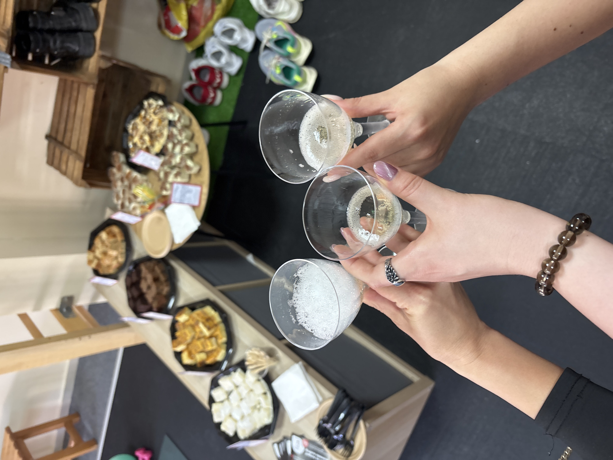 The image depicts an indoor scene with three people toasting with glasses of champagne. The view is from above, focusing on the hands and glasses. A table laden with various food items is visible in the background, including platters of pastries, cheese, and other snacks. Also in the background is a wooden crate with shoes stacked on top. A black surface is seen below the table. The location and city are not identifiable based on the image.