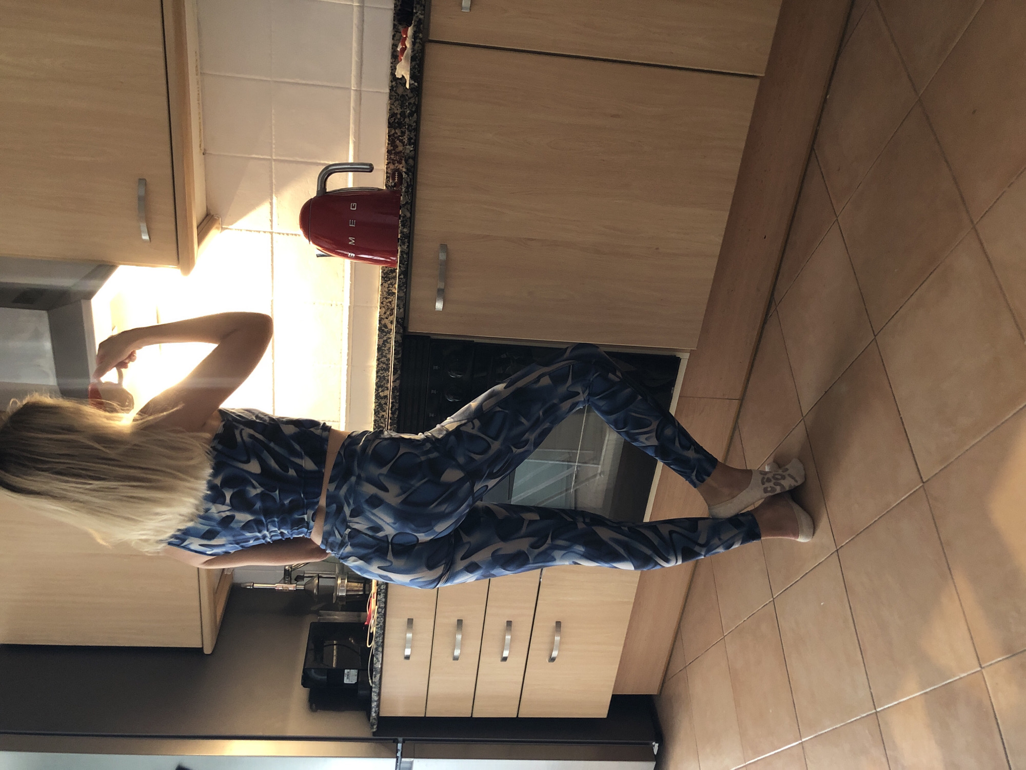 A young woman with blonde hair, wearing a blue and white patterned crop top and matching leggings, stands in a kitchen. She is holding a mug to her lips as she looks up towards a cabinet light. Her feet are positioned with one leg bent and resting on the toes of the other. The kitchen features light-colored wooden cabinets, granite countertops, and tiled flooring. A red Smeg kettle is visible on the counter. The scene appears to be a domestic environment, likely a residential kitchen.