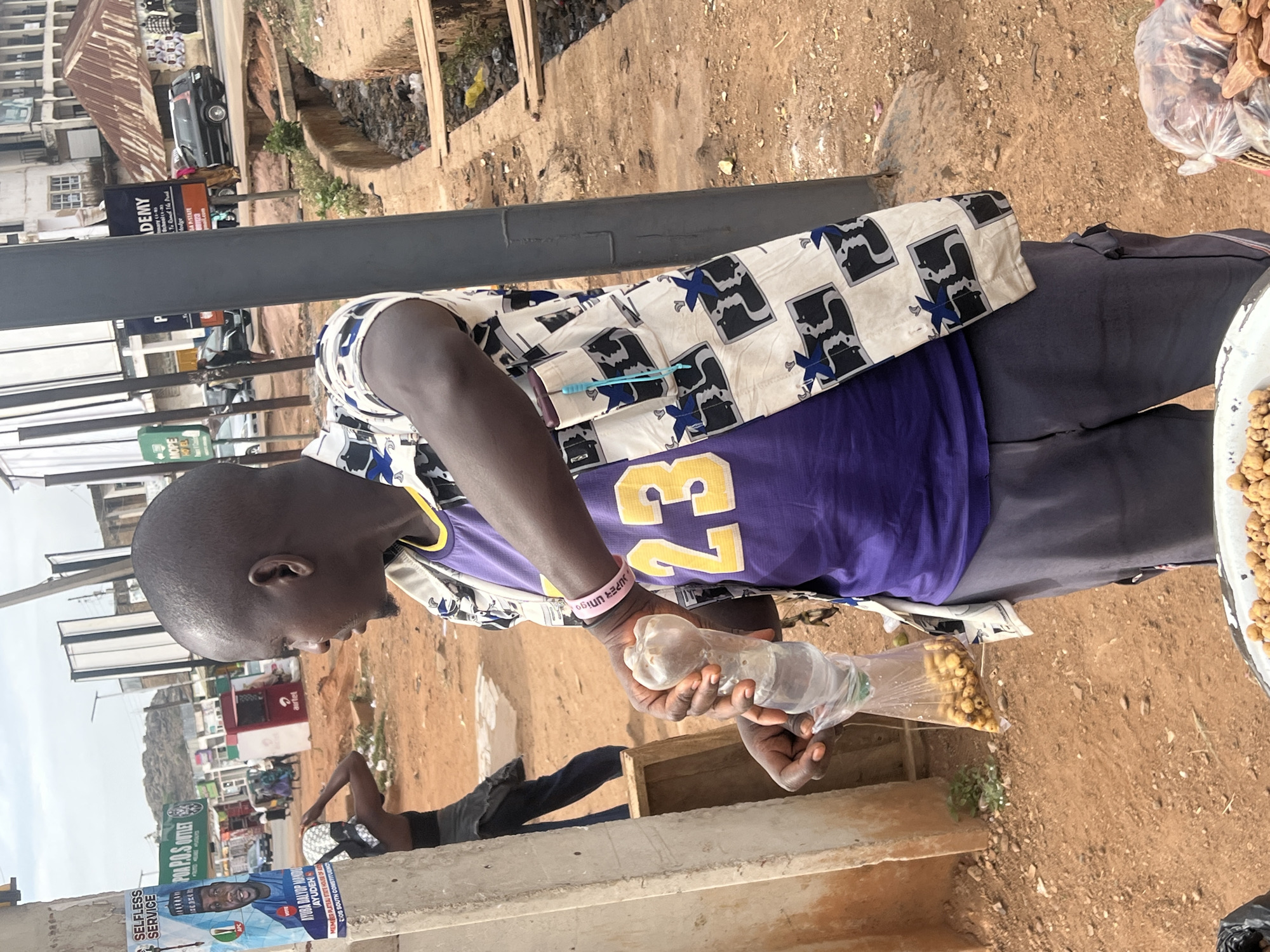 A man wearing a purple Lakers jersey with the number 23 is selling roasted peanuts by the roadside in Jos, Nigeria. He is filling a small plastic bag from a plastic bottle with the peanuts. The environment includes visible city infrastructure such as buildings, utility poles with advertisements, drainage ditches, and roadside vendors. The photograph is taken at a slight angle, showing the ground and immediate surroundings where the transaction is occurring.