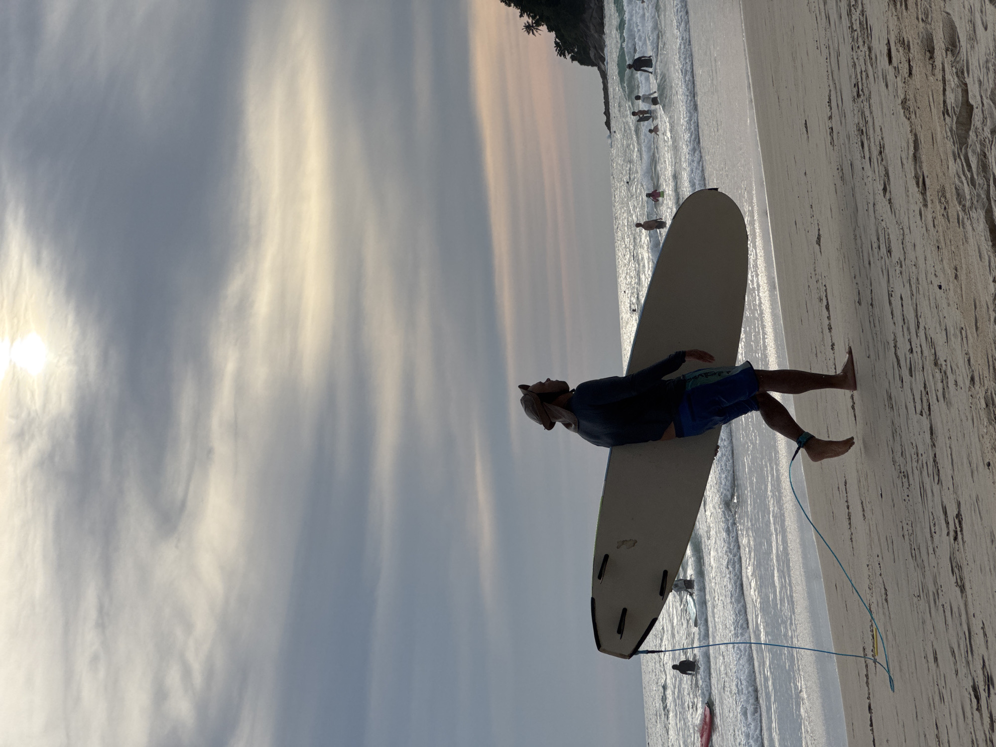 A person walks along the beach in Karon, Thailand, carrying a surfboard. The sky is overcast with diffused sunlight. The ocean has moderate waves, and several people are in the water. The sand is light brown with a few footprints. The person is wearing a hat, a long-sleeved shirt, and blue shorts. A leash attaches the surfboard to their ankle.