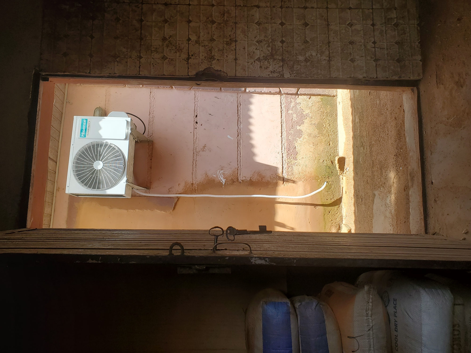 The image shows an outdoor scene, possibly in Bukuru, Nigeria, looking down into an opening. In the upper section of the opening, a white Hisense air conditioning unit is mounted on a pale pink wall. A white pipe runs from the unit and along the wall. Above the air conditioner, the wall is tiled with a geometric pattern.
The opening itself is framed by what appears to be a wooden structure. Below this wooden structure, more of the opening reveals a sandy or dusty area, possibly a yard or a path, with sunlight casting shadows from unseen objects.
On the wooden structure, a dark metal key with a decorative handle is placed. Below the wooden structure, the image shows the tops of several large bags. One bag is white with text printed on it, including "COOL DRY PLACE". Other bags appear to be different colors, one of which has blue material visible.
The lighting suggests it is daytime, with strong sunlight creating distinct shadows. The overall scene appears to be somewhat weathered or aged, with some discoloration and wear visible on the walls and structures. The presence of the air conditioning unit and the bags suggests a functional, perhaps storage or residential, outdoor area.