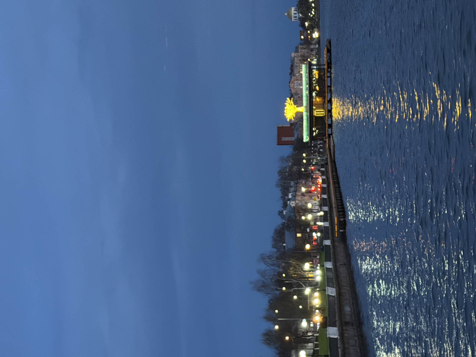 The image displays an evening or nighttime scene of a waterfront in Votkinsk, Russia, under a deep blue sky. The composition, if viewed horizontally, shows a body of water on the right and a city or townscape along the left shore.
The water's surface, likely a lake or river, is dark blue and reflects numerous artificial lights from the shore, creating shimmering streaks of yellow and white light across its expanse. A promenade or pier runs along the water's edge, lined with streetlights that illuminate the path and surrounding areas.
On the land side, beyond the promenade, the outlines of bare trees are visible, suggesting a winter or early spring season. Various buildings form the cityscape in the background. A distinctive golden-domed building, possibly a church or public edifice, is visible in the distance. Closer to the foreground, a prominent reddish-brown building stands out. A striking focal point is a large, brightly illuminated yellow sculpture, resembling an abstract tree or starburst, situated on the promenade near the water. This sculpture adds a decorative and vibrant element to the scene. Additional lights from distant buildings and what appear to be street or vehicle lights dot the urban landscape.
The scene conveys a peaceful and atmospheric ambiance, characteristic of the "blue hour" or early night, with the artificial lights providing warmth against the cool tones of the sky and water. No specific activities or people are visible, nor is any text discernible in the image.