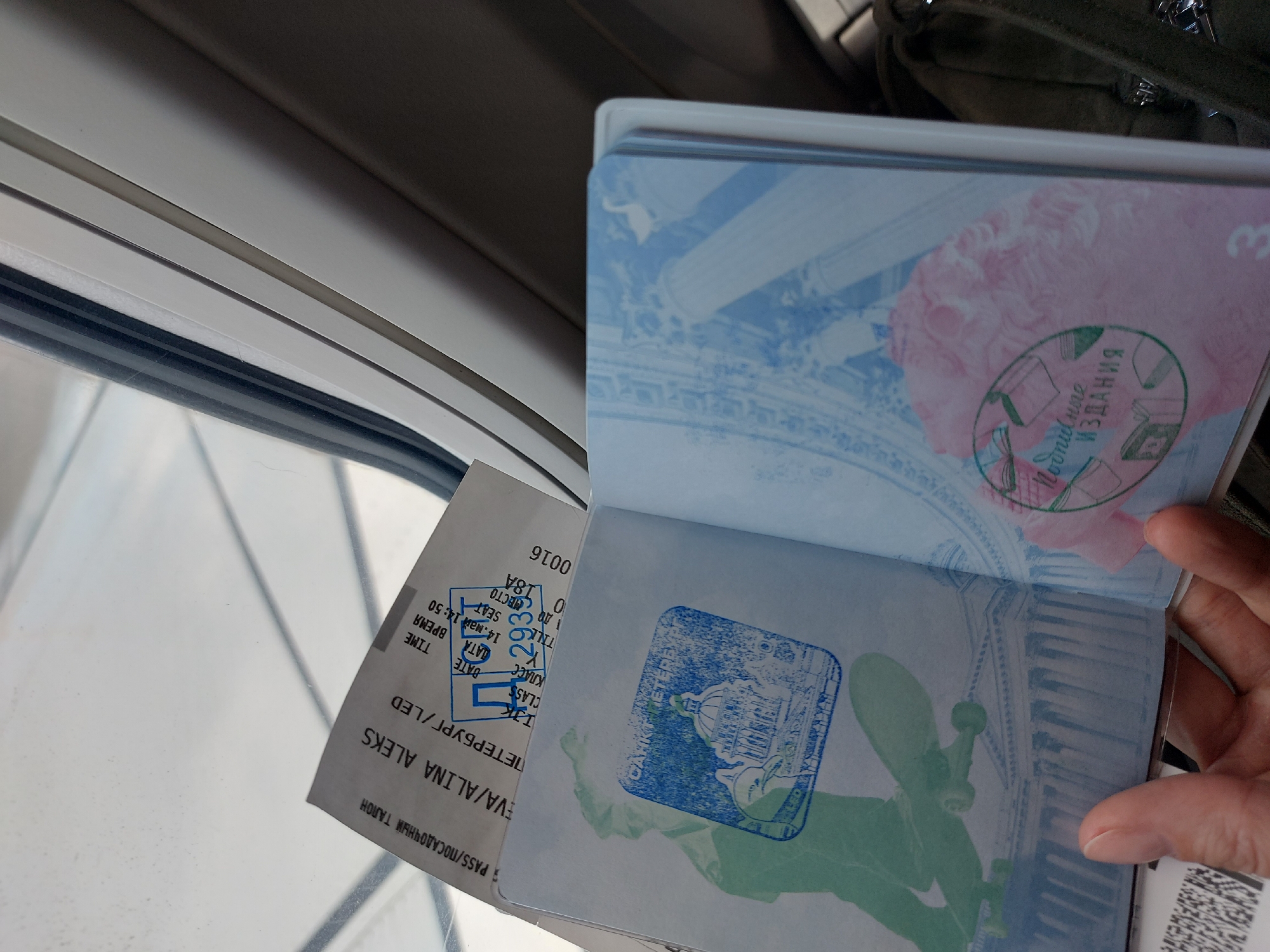 The image shows a passport opened to reveal Russian visas. A boarding pass is placed next to the passport. The boarding pass is for a flight from Saint Petersburg (LED) to an unknown destination. The passenger's name is EVA/ALINA ALEKS. The boarding pass indicates a flight on May 14 at 14:50. The photo appears to have been taken in an airplane, as a window and seat are visible. One of the visa pages features a blue-green image of the statue of Apollo at the Saint Petersburg Mining University. Another visa page features a green stamp. The person holding the passport has fair skin. A backpack is slightly visible in the background.