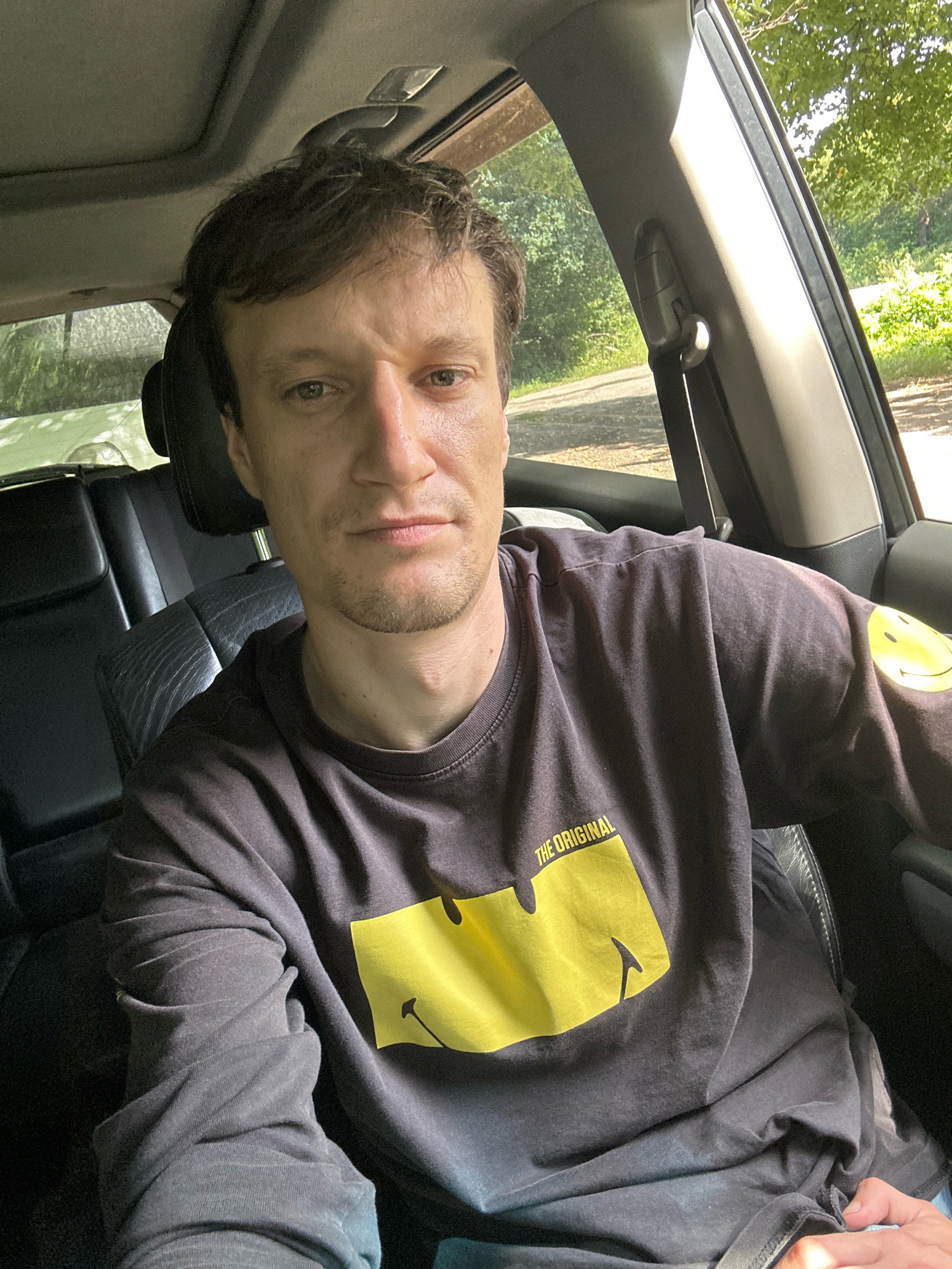 A man is sitting in the driver's seat of a car, looking at the camera. The car's interior is visible, with black leather seats and a sunroof. The man is wearing a dark grey long-sleeved t-shirt with a yellow smiley face graphic and the words "THE ORIGINAL" printed on the front. His right sleeve also has a yellow smiley face. The background outside the car shows trees and foliage, indicating an outdoor setting. The image does not provide any specific information about the city of Shatava, Ukraine.