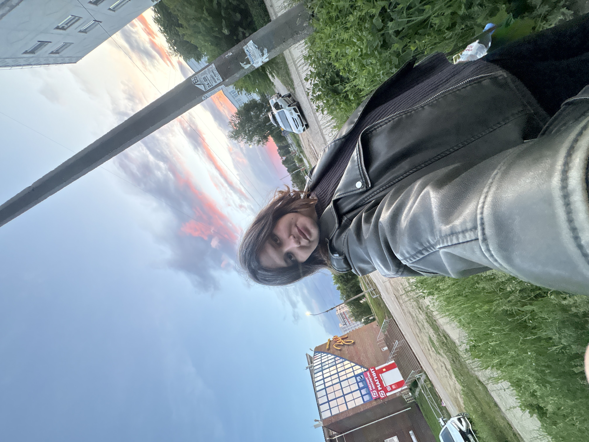 A selfie photograph depicts a woman wearing a leather jacket and a black top. A water bottle is visible inside her jacket. The background shows a street scene with a building labeled "МАГНИТ" (Magnit, a Russian retail chain) and other structures. Cars are parked along the street. The sky is visible with colorful clouds during sunset. The location is Chaikovsky, Russia, as indicated by the "МАГНИТ" retail chain and other visual elements common in Russian urban settings.