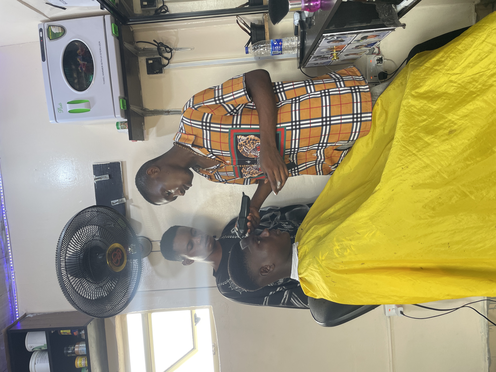 Inside a barber shop in Ado, Nigeria, a barber in an orange plaid shirt is giving a haircut to a customer draped in a yellow cape. Another man watches the barber. A black electric fan stands nearby. A towel warmer with UV sterilizer is mounted on the wall. Bottles and containers are visible on a shelf.
