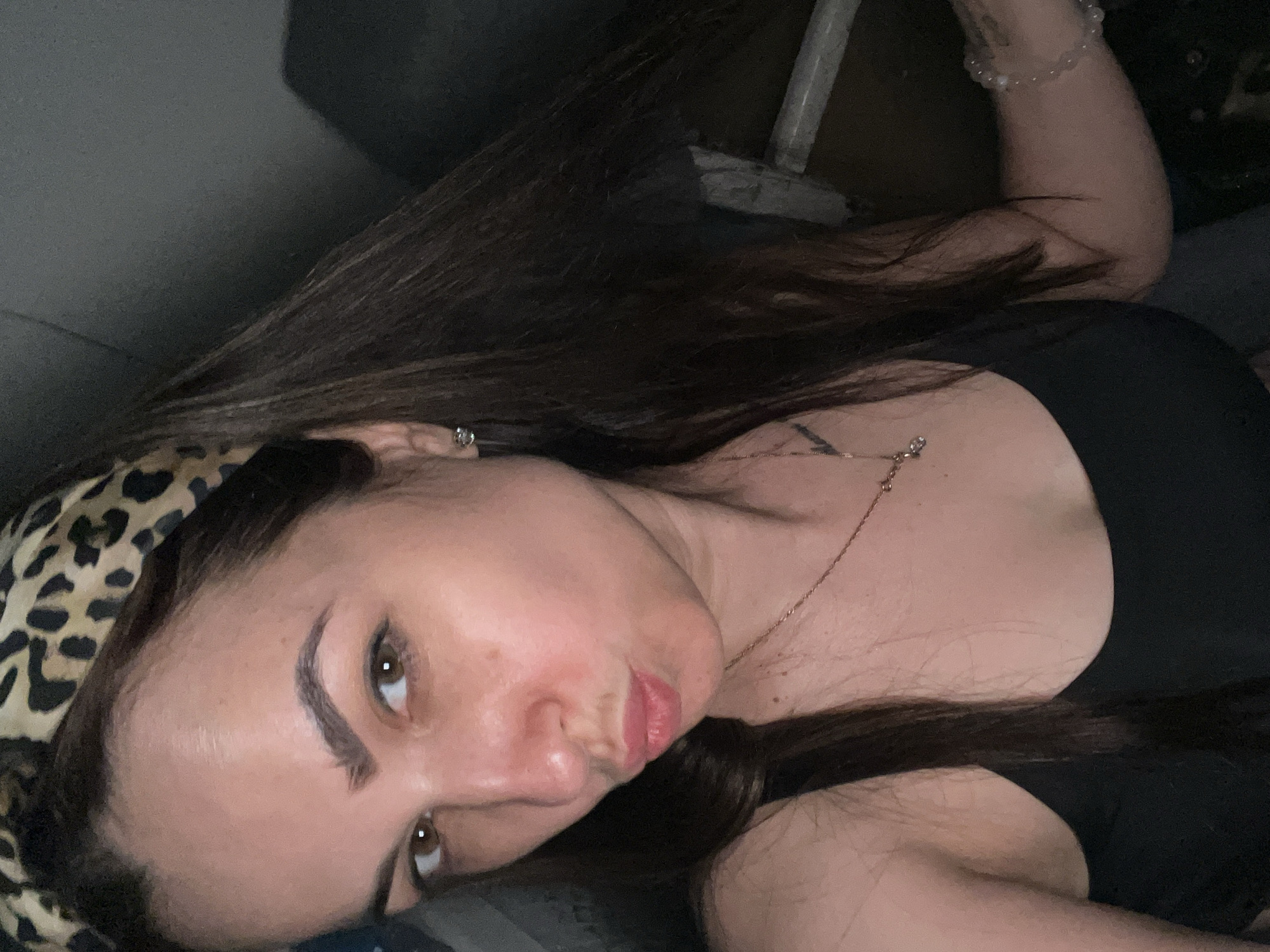 A woman lies on her side, facing the camera. She is wearing a leopard-print headband and a black top. She has long, dark hair. A necklace is visible around her neck. Her right arm is bent, and a beaded bracelet is on her wrist. The background is dark and unclear.