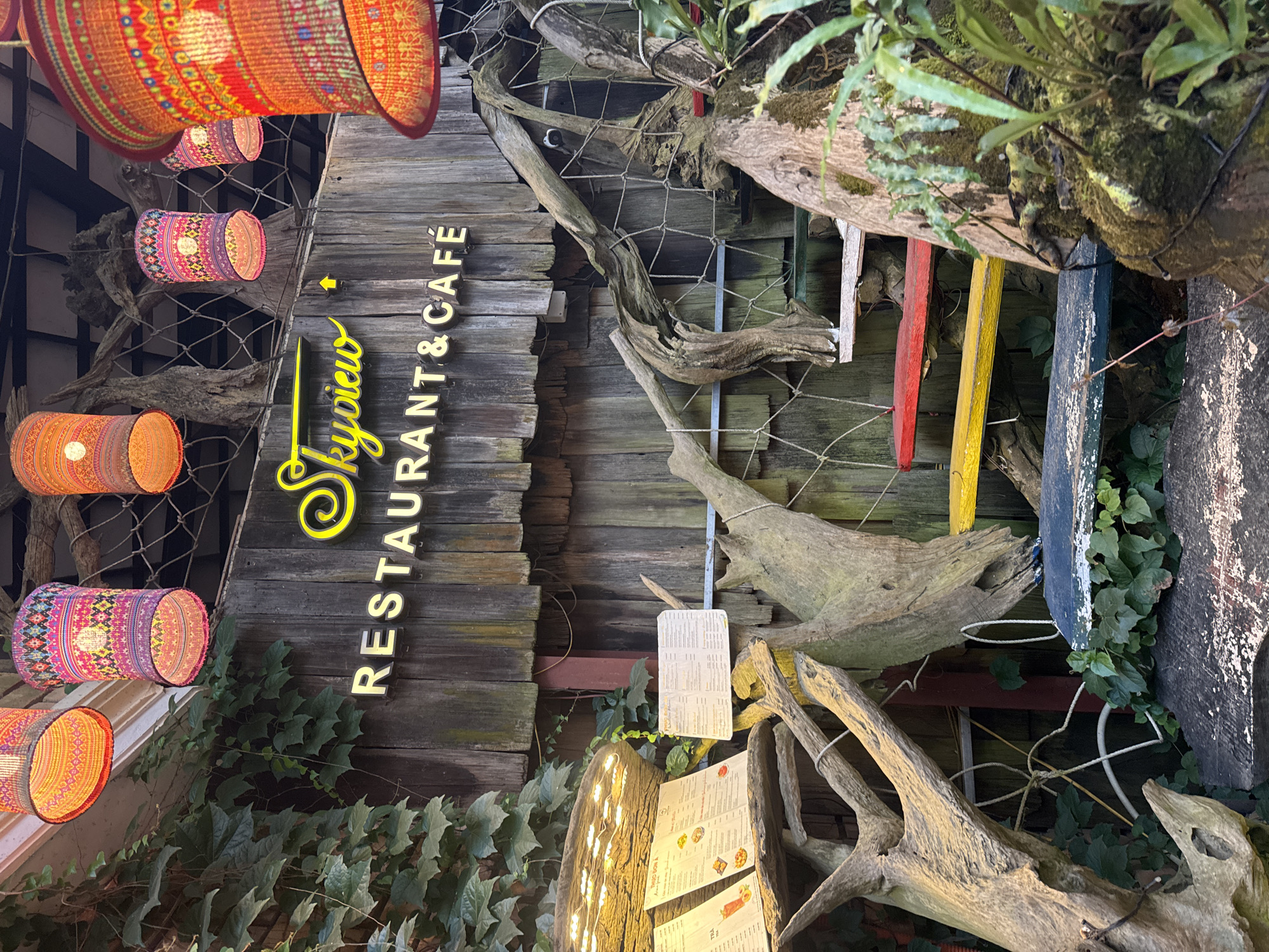 Captured in Sa Pa Ward, Vietnam, this image presents the exterior of "Skyview Restaurant & Cafe." The facade features rustic wooden planks, adorned with the restaurant's illuminated signage. Decorative cylindrical lanterns hang overhead, adding a touch of vibrancy. The architecture is intertwined with natural elements: gnarled tree branches, greenery, and hanging moss, creating a unique aesthetic. Restaurant menus are displayed, suggesting a casual dining experience.