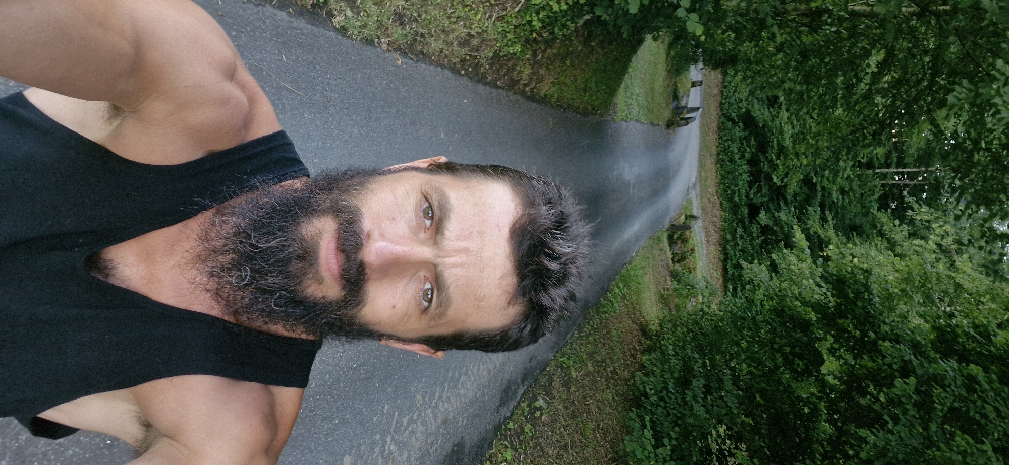 The media file is a self-portrait of a man with a full beard, taken outdoors. The man is wearing a black tank top. He appears to be in good physical condition, with visible muscles in his arm and shoulder. His expression is neutral, with his eyes looking directly at the camera.
The scene is a paved path, likely a trail or road, surrounded by lush green trees and foliage. The path appears wet, suggesting recent rain or damp conditions. There are some stones or low benches visible along the side of the path, and in the distance, two figures can be faintly seen walking away.
The lighting suggests it is daytime, possibly overcast or late afternoon, given the soft light and lack of harsh shadows. The overall mood is serene and natural, with the focus on the man and his surroundings. There is no visible text in the image. The image is oriented vertically, with the man's head towards the right side of the frame.