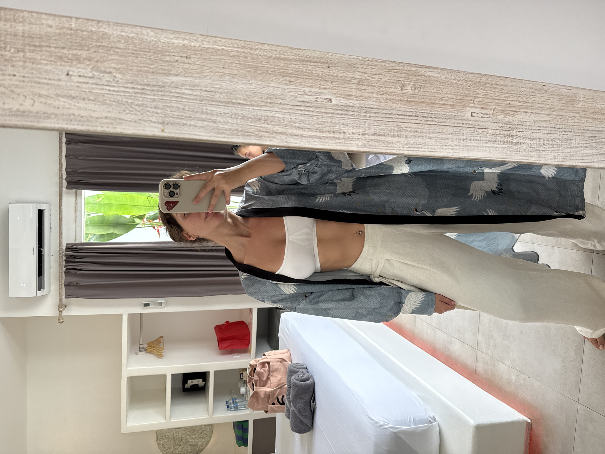 A medium shot taken indoors in a location determined to be in Unknown, Indonesia depicts a Caucasian person taking a mirror selfie with an iPhone. The person is wearing a white bra top and light colored loose-fitting pants with a light blue kimono patterned with white birds. The person is standing in what appears to be a bedroom. Background objects include, an air conditioning unit mounted on the wall near a window with partially closed blinds, a white shelving unit with various items, including a red bag and bottled water, and the corner of a white bed. The setting is clean and well-lit. The image is slightly out of focus due to being a photograph taken in a mirror.