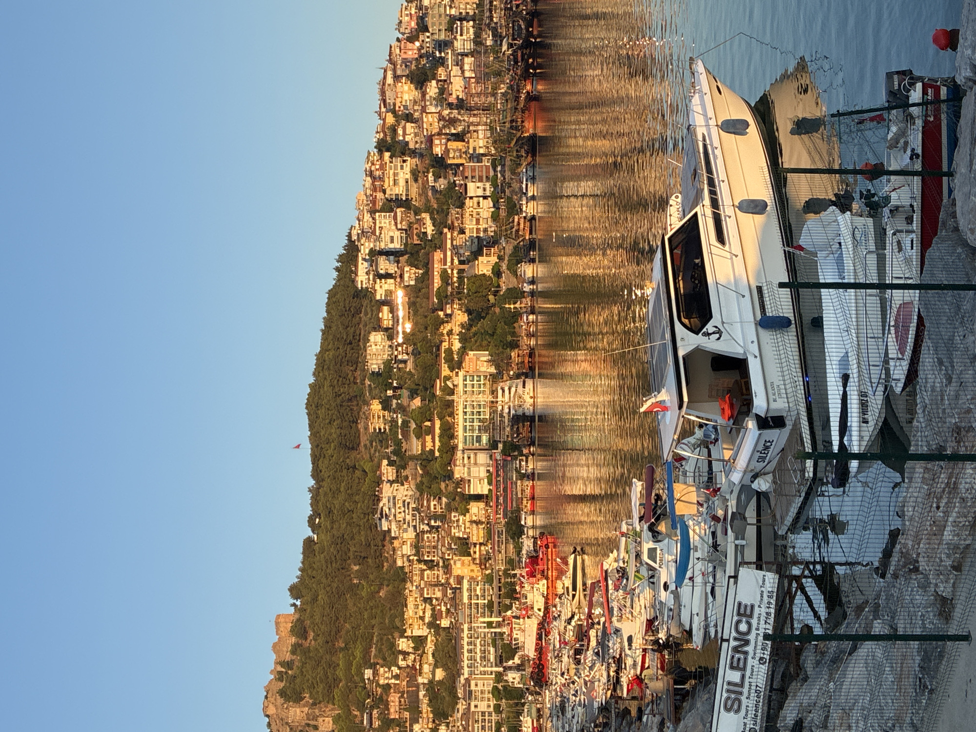 The image depicts a harbor in Alanya, Turkey, with a hillside city overlooking the water. The sky is clear and blue, indicating daytime. Numerous boats are docked in the harbor, ranging from small personal watercraft to larger vessels. Some boats are white, while others are colorful, adding to the visual appeal of the scene. The buildings on the hillside are a mix of colors and architectural styles, with many featuring terracotta roofs. A prominent castle ruin sits atop the hill, suggesting historical significance. The water reflects the sunlight and the surrounding scenery, creating a shimmering effect. The overall atmosphere is peaceful and serene, with the calm waters and warm sunlight contributing to a sense of tranquility.