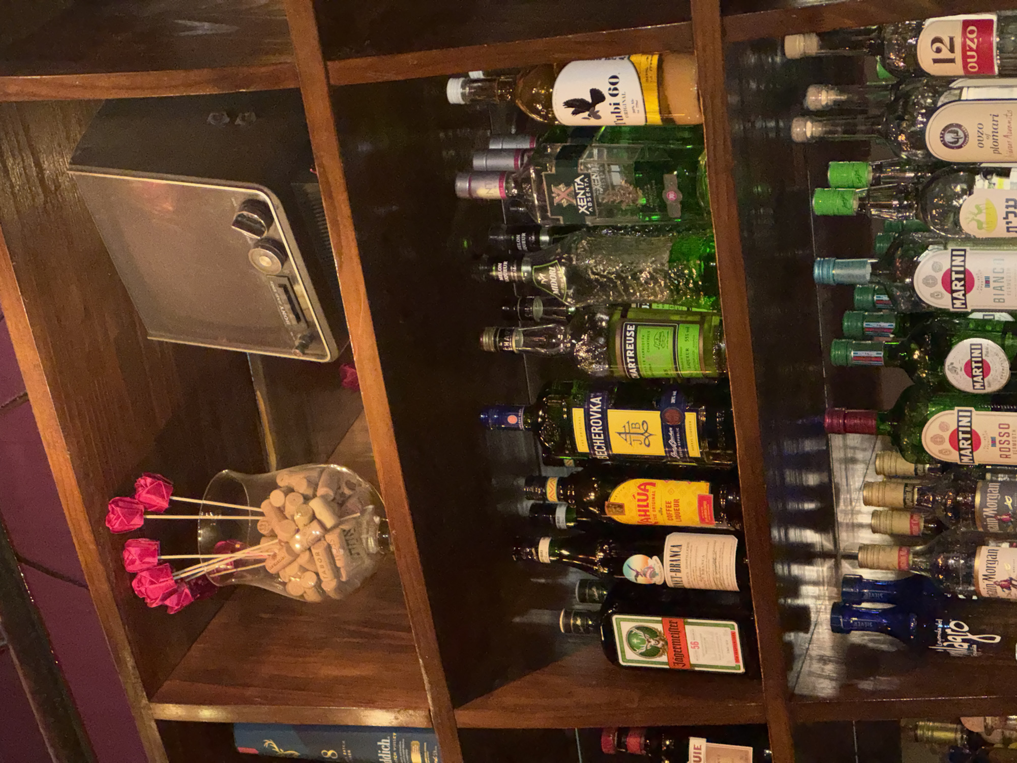 The image displays three dark-stained wooden shelves stacked with various bottles and decorative items, indicative of a bar or pub interior.
On the top shelf, a clear glass vase holds multiple wine corks and five artificial pink tulip-shaped flowers on slender stems. To its right, a dark gray vintage Sony television or monitor is positioned, featuring a silver trim and two circular control knobs on the right side of its screen. A partially visible dark blue object on the far left, possibly a book or bottle box, bears the text "8" and "GLENFIDDICH."
The middle shelf showcases an array of liquor bottles. From left to right, identifiable brands include Jägermeister, Fernet-Branca, Kahlúa Coffee Liqueur, Becherovka, Chartreuse, Midori Melon Liqueur, Xenta Absenta, and Tubi 60, which has a white label and a black bird silhouette.
The bottom shelf also contains numerous liquor bottles, including Legenda del Milagro (in blue bottles), Captain Morgan, Martini Rosso, Martini Bianco, a clear bottle with Hebrew script (likely an Arak brand), Ouzo of Plomari, and 12 Ouzo.
The background consists of a dark wall, with a purplish hue visible in the upper left. No individuals are present in the scene. The inclusion of Tubi 60, a well-known Israeli beverage, and a bottle with Hebrew text strongly suggests the establishment is located in Israel. While a precise address in Tel-Aviv is not identifiable from the image, the depicted liquor selection is consistent with a bar in that city.