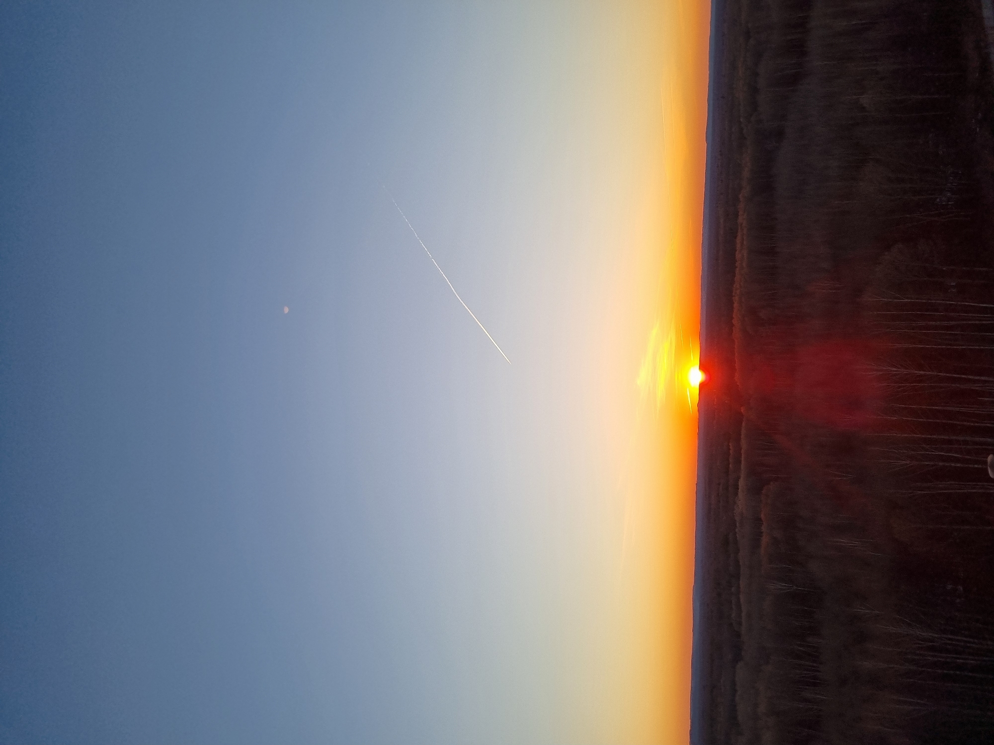 The image shows a sunset over a dark forest in what is likely Kemul, Russia. The sky transitions from blue to orange near the horizon. A contrail from an aircraft stretches across the sky. A faint object resembling a moon is visible in the upper left. The sun is partially obscured by the forest, creating a bright orange glow with a red flare around its edge.