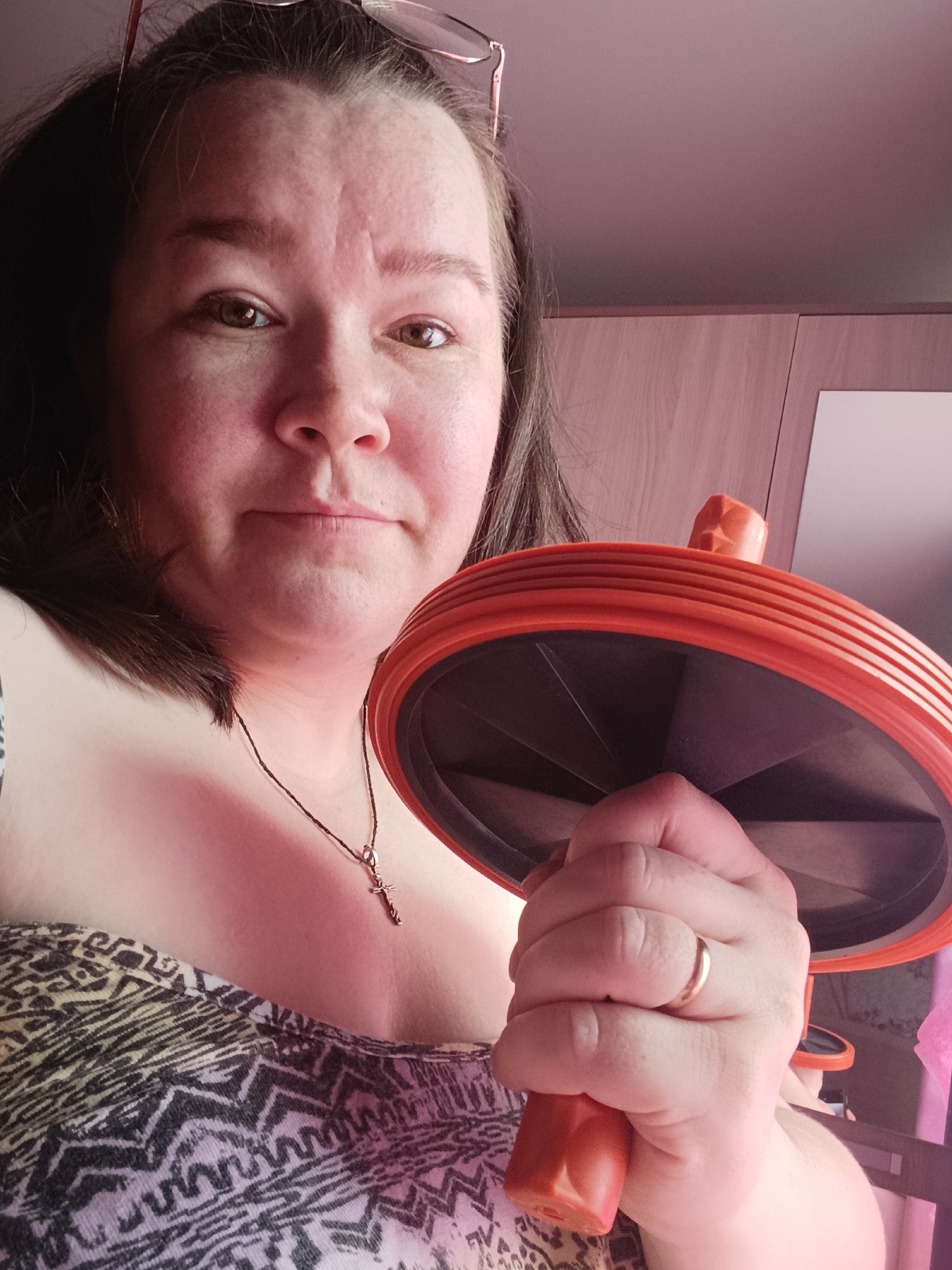 An adult woman is featured in an eye-level, close-up portrait, holding an orange and black ab roller in her right hand. She has dark hair, which is pushed back with a pair of glasses resting on top of her head. Her brown eyes look directly at the camera, and she has a neutral, composed expression. She is wearing a patterned, possibly strapless or off-the-shoulder top with a black, white, and gray geometric design. A necklace with a small silver cross pendant hangs around her neck, and a plain gold band is visible on her left ring finger. The background consists of wooden cabinets or furniture, indicating an indoor setting. The lighting is soft and diffuse, suggesting indoor illumination rather than direct sunlight. The scene takes place in Chaikovsky, Russia, and no text is visible in the image.