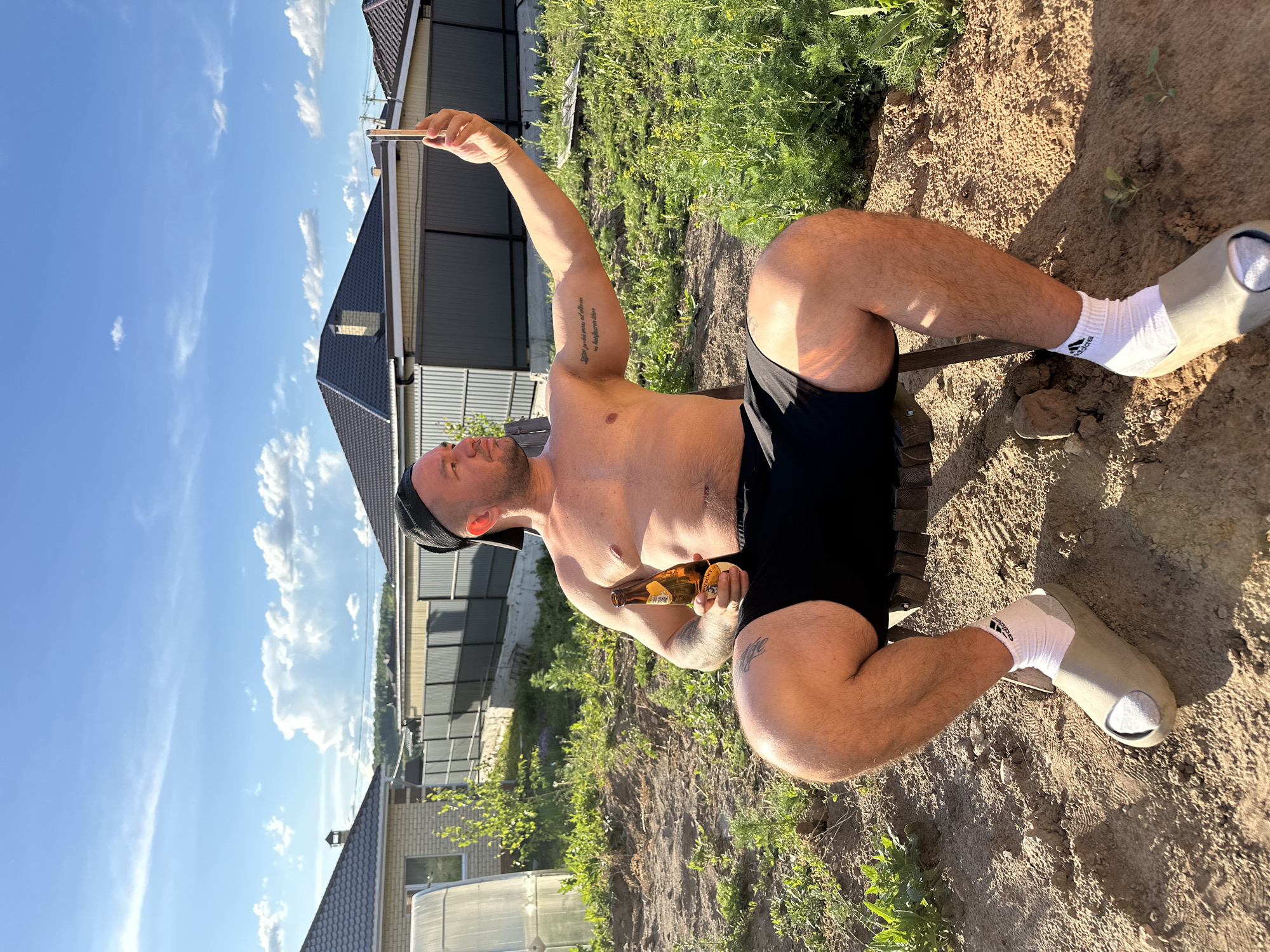 A man is sitting on the ground holding a cell phone in his raised right hand and a beer bottle in his left hand. He is shirtless and wearing black shorts, white socks, and beige sandals. He is also wearing a black cap. The background includes a house with a gray roof, green grass and plants, a greenhouse, and a blue sky with white clouds. The ground appears to be dirt or sand. The location is not specified.