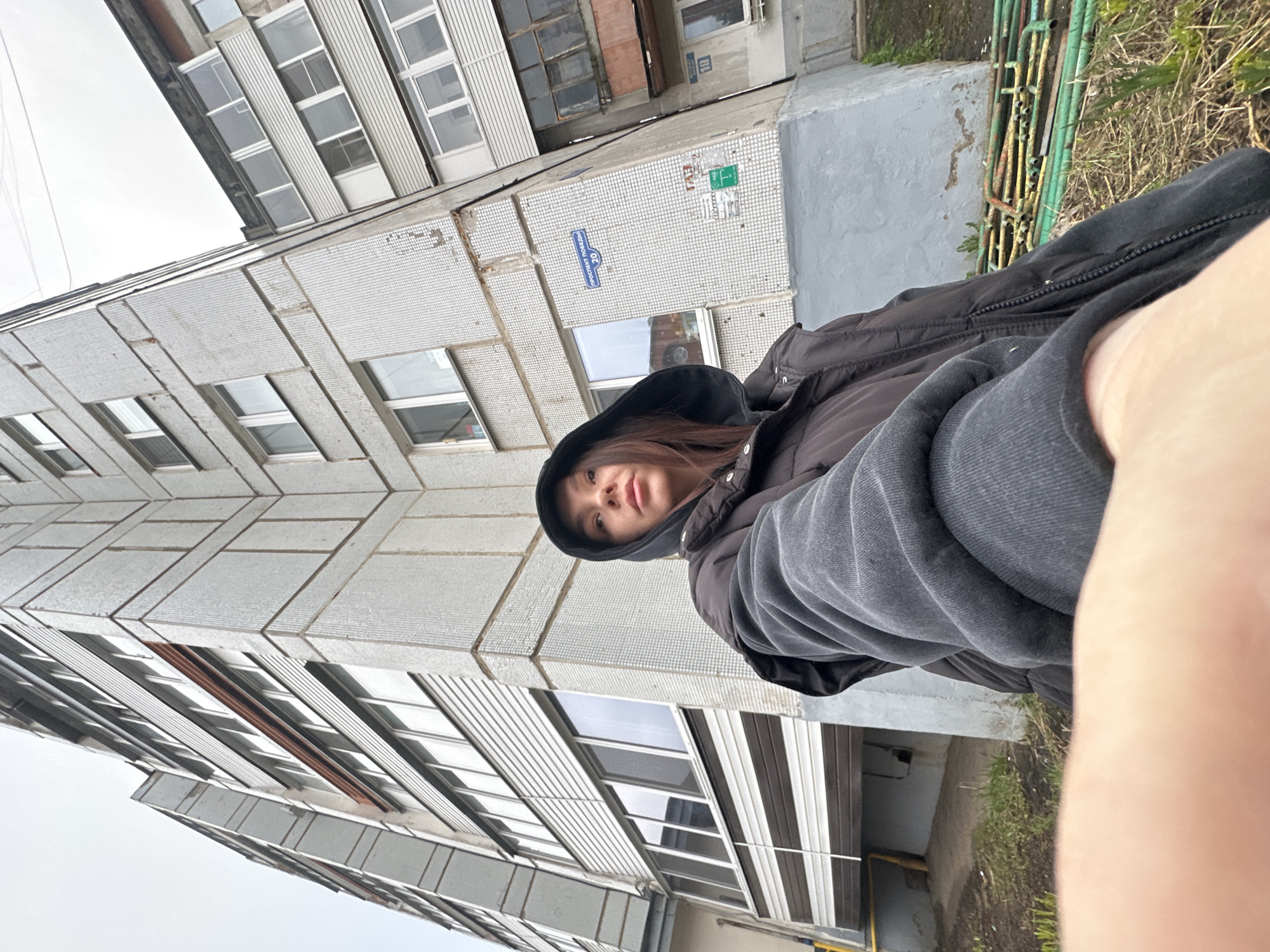 A selfie taken by a young woman wearing a black hooded jacket and a grey sweater. She is standing outside a grey, tiled building with multiple rows of windows. The building appears to be an apartment complex. There are green metal pipes near the building and some grass. A street sign, partially obscured, includes Cyrillic script indicating a location in Chaikovsky, Russia. The photo is taken from a low angle, looking up at the building. Part of the photographer's arm is visible on the right side of the frame.