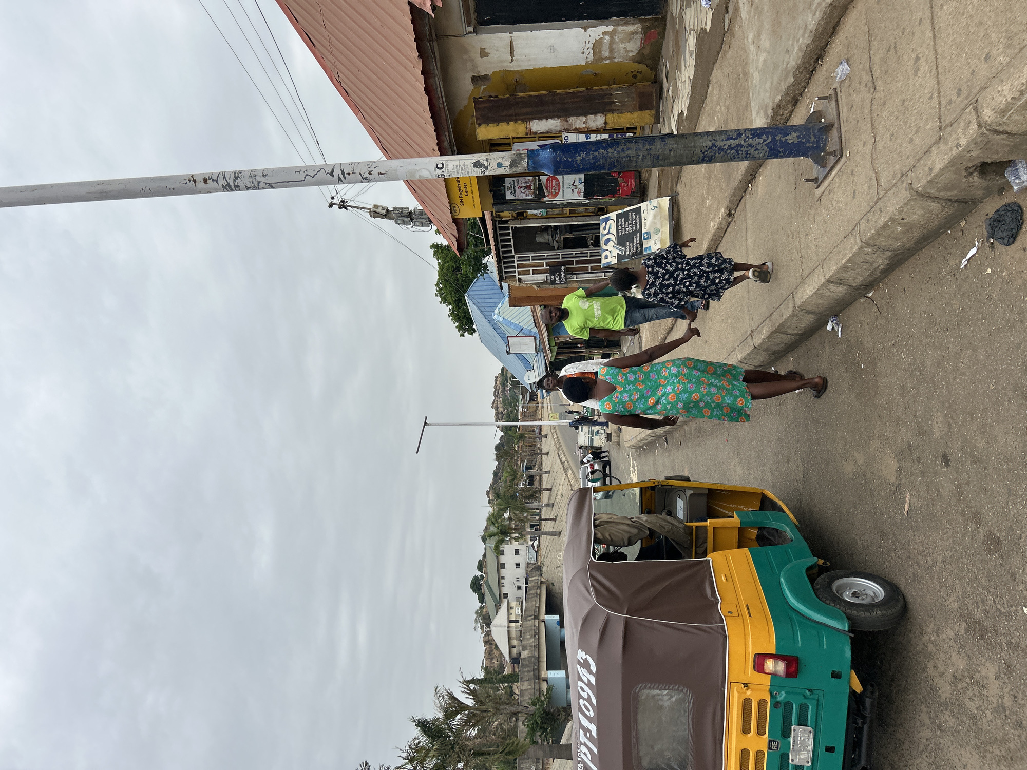 The scene depicts a street in Jos, Nigeria. A three-wheeled vehicle, predominantly green and yellow, with a dark brown cover, is parked on the right side of the street. The vehicle's license plate is visible and displays "PT-10993".
Three individuals are walking along the sidewalk adjacent to the vehicle. The person closest to the vehicle is wearing a green floral print dress. The second person is wearing a bright green shirt. The third person is wearing a dark-colored, patterned dress.
Adjacent to the sidewalk is a building with a red roof. A sign that reads "SIM Registration Center" is visible. Another sign reads "POS", with additional text below.
Across the street, palm trees and buildings are visible.