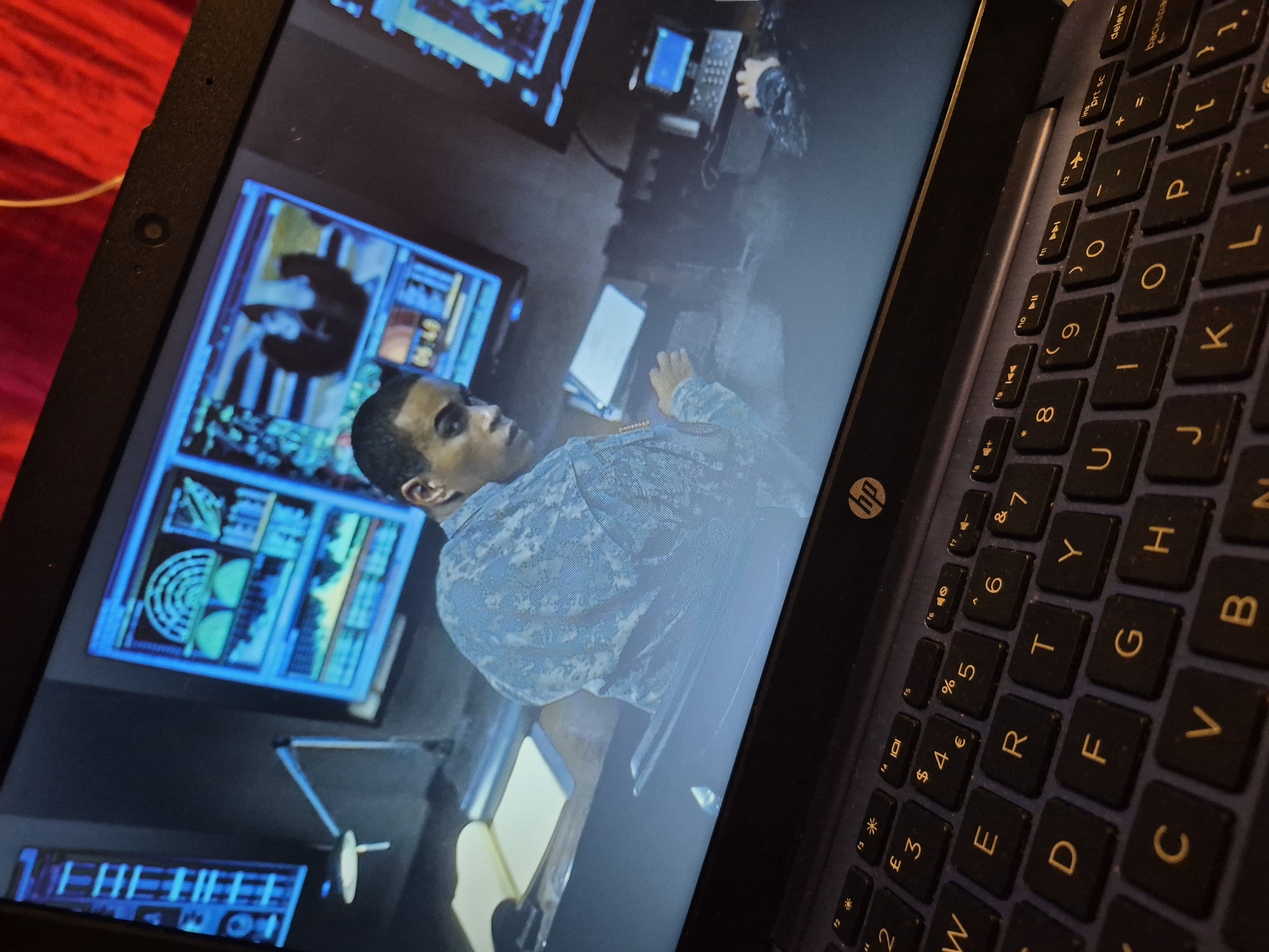 An individual wearing a light blue patterned military-style uniform is seated in front of a laptop, their gaze directed towards the screen. The laptop displays a scene featuring multiple glowing blue computer monitors, one of which shows a person. The environment appears to be a dimly lit room with a desk that has a document and a lamp on it. The laptop's keyboard, with the HP logo visible, occupies the foreground. The image does not provide any information about the city of Sandwell, United Kingdom, or any specific location within it.