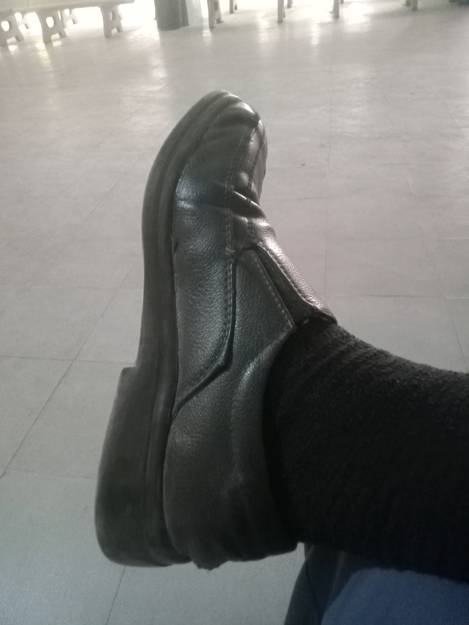 A close-up, low-angle shot shows a person's left foot wearing a black leather loafer and a black sock. The shoe has a thick, dark sole and a smooth, pebbled leather upper. The sock appears to be ribbed. The foot is resting on a tiled floor, which is light gray with faint grid lines. In the background, out of focus, are white tables or benches. The lighting suggests an indoor environment, possibly a waiting area or common space. The image is taken from a perspective that focuses on the foot and shoe. No specific location in Karachi Division, Pakistan can be identified from the visual elements alone.