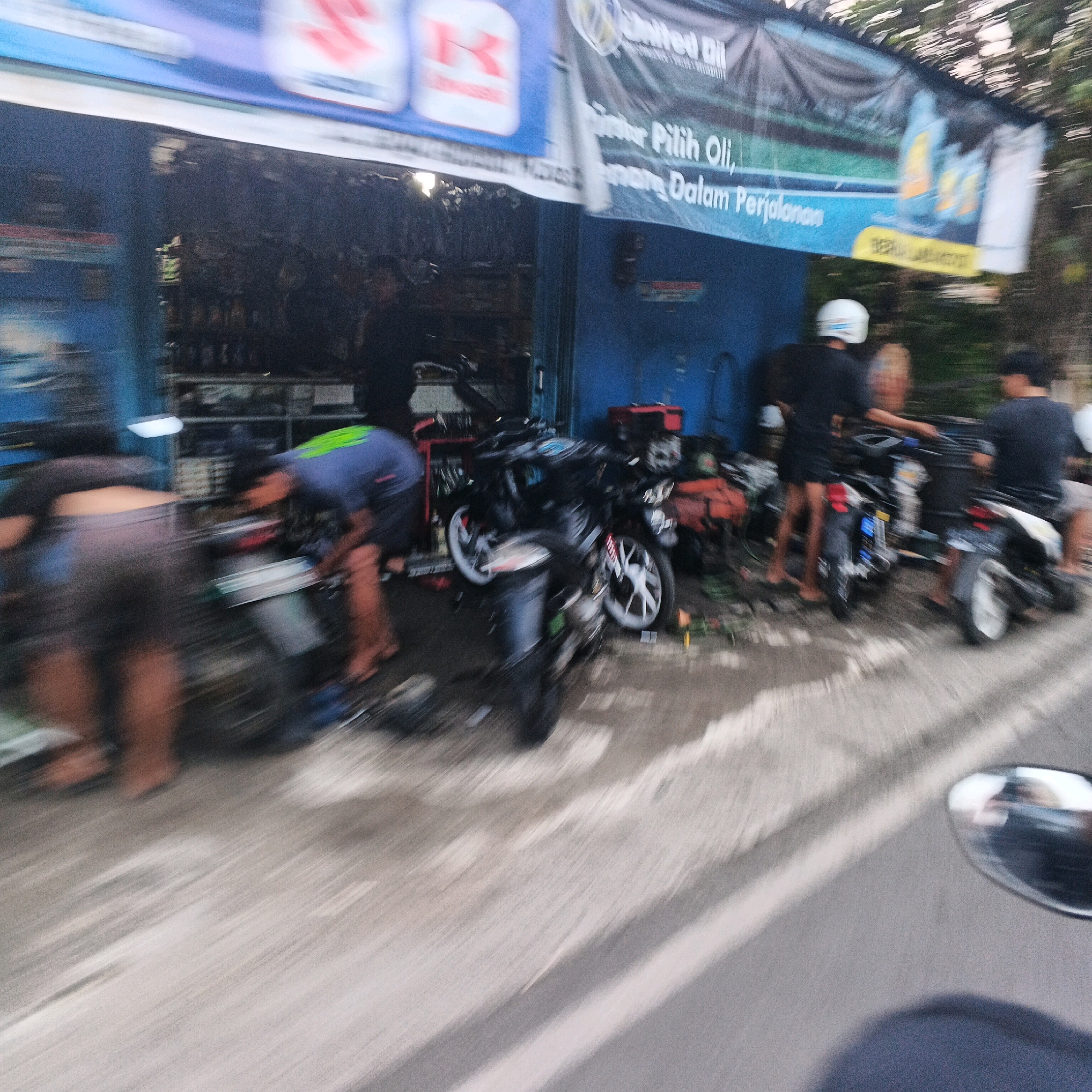 In Kalikudi, Indonesia, a blurry, motion-captured image depicts a roadside motorcycle repair shop. Two individuals are visible, one in the foreground wearing a blue shirt with green accents, bent over a black motorcycle. Another person in the background wears a black shirt and a white helmet. Several motorcycles are parked outside the open-fronted shop, which has a blue wall and shelving stocked with items. A banner overhead, bearing the "United Oil" logo, is also visible, along with other banners and signage in Indonesian. The scene appears to be an active workshop with customers or workers present.