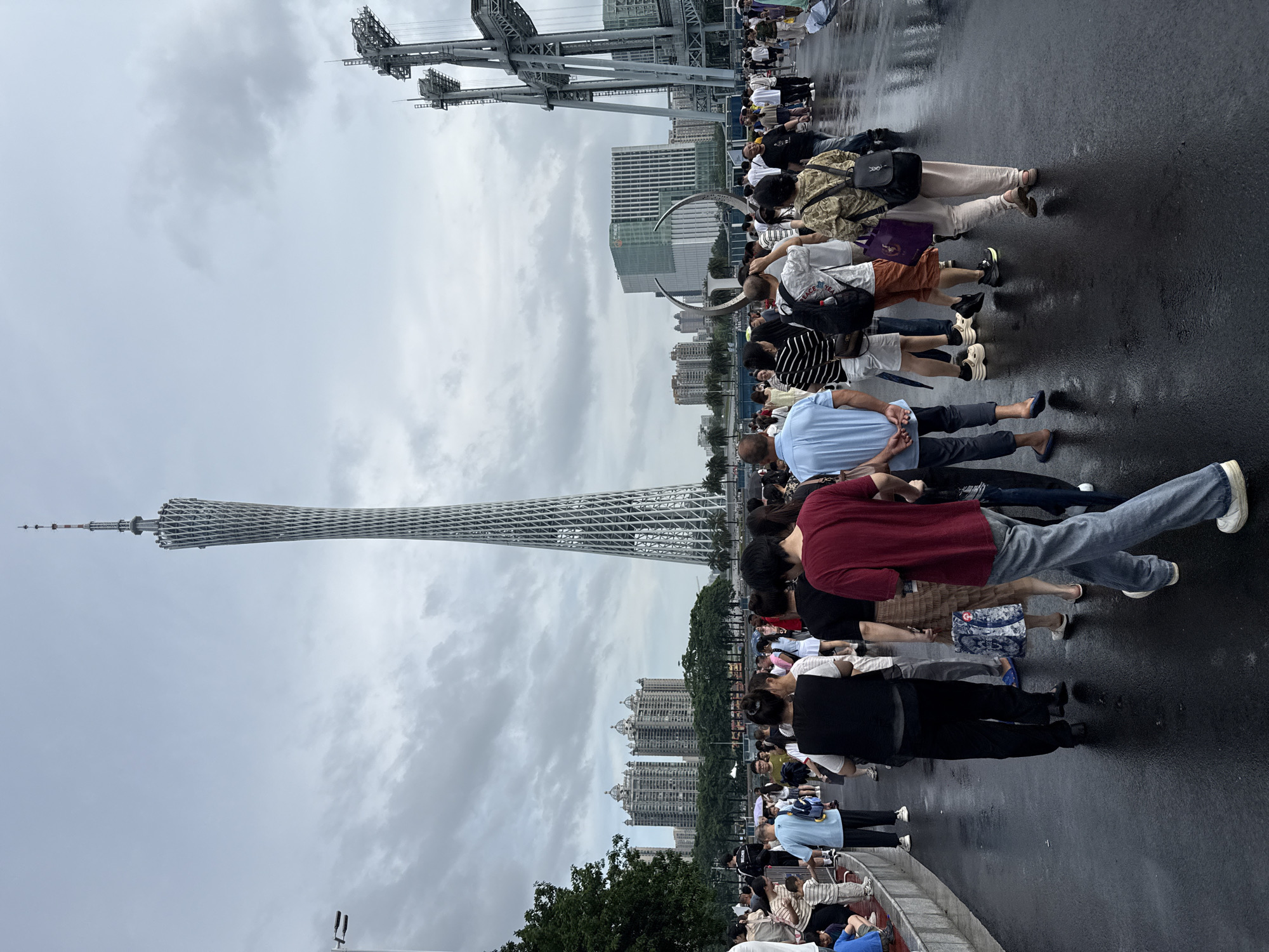 The image captures a crowd of people walking on a wet, paved area with reflections visible on the surface, suggesting recent rain. In the background, the Canton Tower, a prominent landmark in Guangzhou, China, stands tall, characterized by its distinctive lattice design. To the right of the tower, a large, grey, industrial-looking structure with multiple elevated platforms and cables is visible, possibly part of a bridge or transportation system. Further in the distance, various buildings, including apartment complexes with domed roofs and modern office buildings, punctuate the skyline under a cloudy sky. The scene appears to be a public thoroughfare or waterfront area in Tianhe District, Guangzhou, China, with people milling about and heading in various directions. Some individuals are carrying bags and umbrellas, further indicating the recent rainfall.