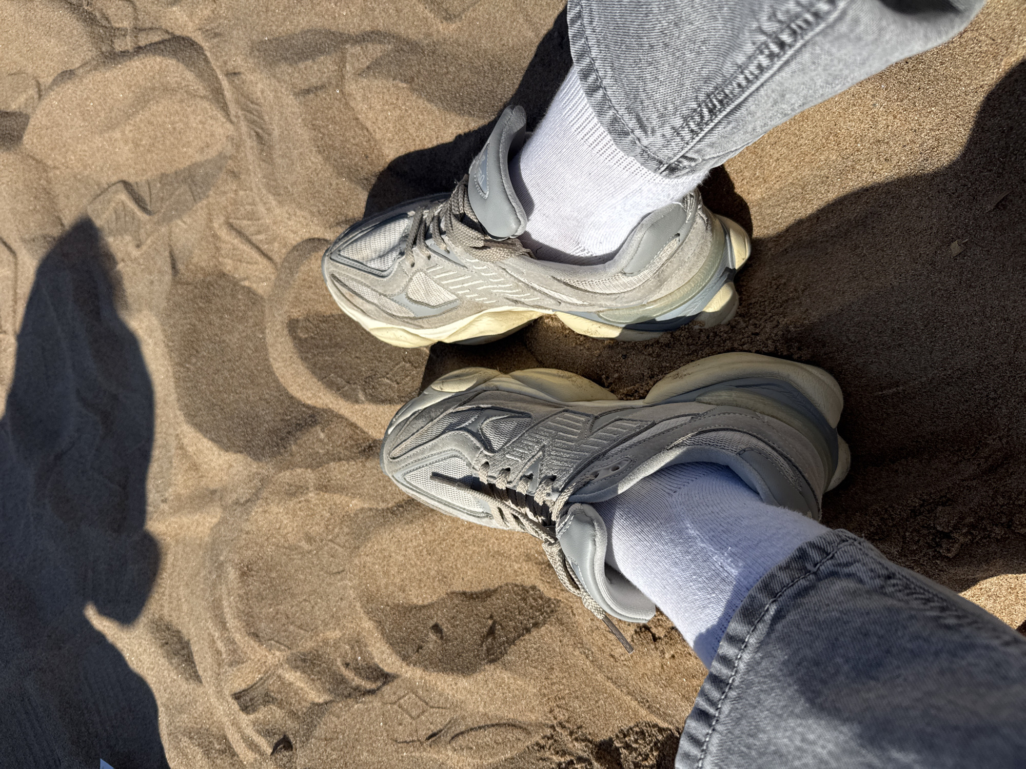 The image depicts a pair of feet wearing grey sneakers, white socks, and grey jeans standing on a sandy surface. The sand is light brown and shows patterns created by wind or other disturbances. The location is Komarovo, Russia.