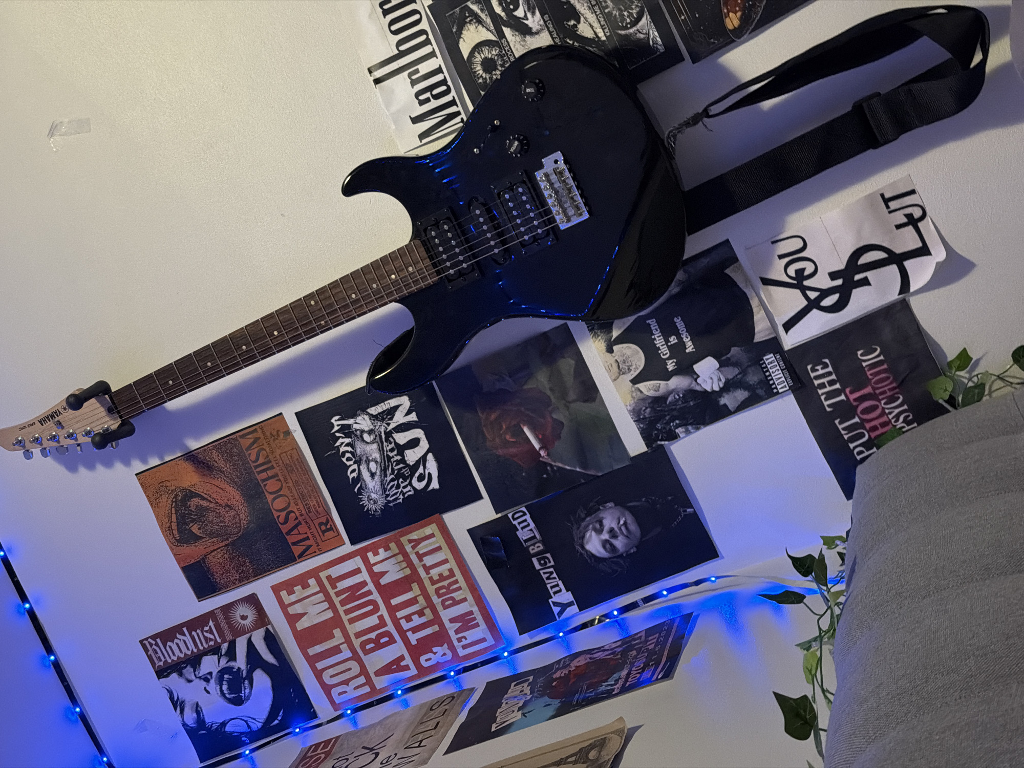 The image depicts an indoor scene with a black electric guitar hanging on a wall adorned with various posters and string lights. The posters feature a mix of graphic art, text, and imagery, including what appears to be album art or band posters. The overall aesthetic suggests a youth-oriented or alternative subculture. The scene does not provide any specific visual cues to indicate its location within the city of Oslo, Norway. There are no people visible in the image, and the objects are static. The environment appears to be a room, likely a bedroom or a practice space, with a textured wall surface and a piece of furniture, possibly a sofa, in the foreground. The lighting is a mix of natural light and the blue glow from the string lights.