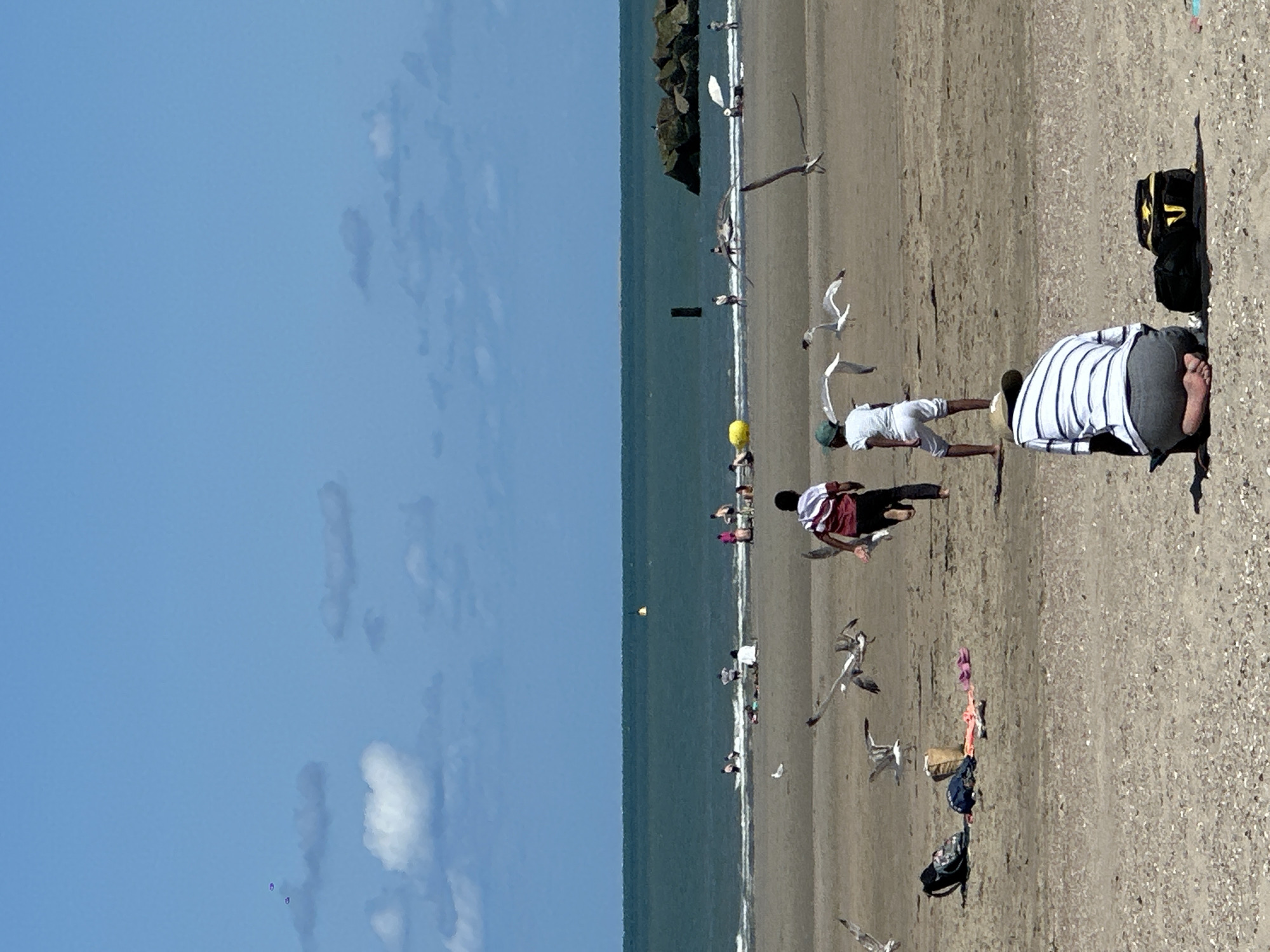 This is a horizontal, eye-level, full shot of a sandy beach in Dunkirk, France, on a sunny day with a bright blue sky dotted with white clouds. In the foreground, a person wearing a striped shirt and gray shorts is kneeling on the sand, facing away from the camera. Next to them is a black bag and a yellow and black bag. In the middle ground, two children are running towards the water, and several seagulls are flying around them. Further back, other beachgoers are visible wading in the shallow water, sitting on the sand, and playing. The ocean stretches across the horizon, and a rocky breakwater juts out into the sea on the right side of the frame. A yellow buoy is visible in the water.