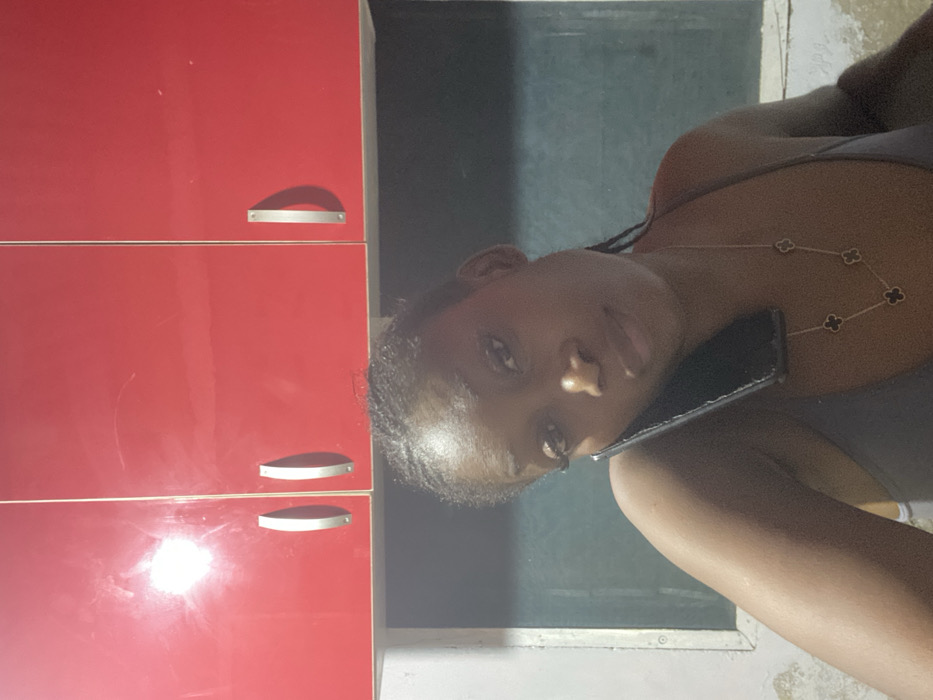 A woman in a gray tank top poses with a cell phone. She wears a necklace with clover-shaped pendants. A red kitchen cabinet with rounded metal handles is positioned beside her. Reflected light creates a bright spot on the cabinet. The background includes a window and light-colored walls. The image is slightly rotated, causing a tilted perspective.