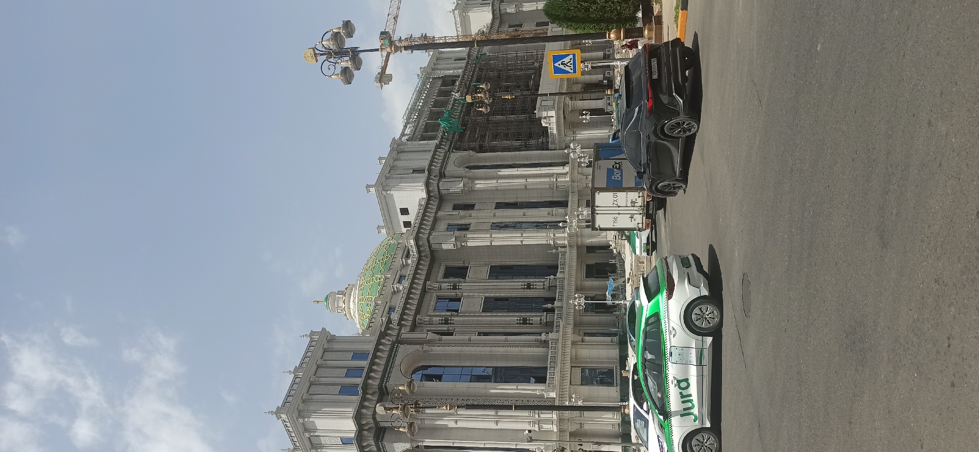 A wide-angle shot captures the facade of a large, ornate building with a green dome, a construction crane, and traffic on a street in Dushanbe, Tajikistan. The building, likely under renovation or construction, displays classical architectural elements like columns and decorative carvings. Scaffolding is visible on the right side of the building, indicating ongoing work. In the foreground, several cars, including a white taxi with the logo "Jura" and a black SUV, are on the asphalt road. A pedestrian crossing sign is present on the right side of the street. The sky is partly cloudy, suggesting a daytime scene.