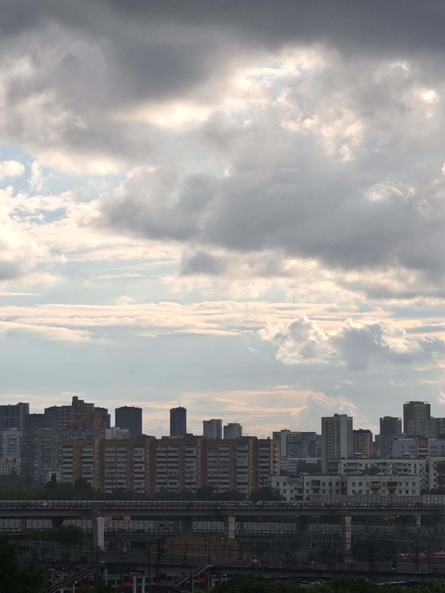 The image depicts a cityscape under a cloudy sky, likely in the late afternoon or early evening given the lighting. The foreground features a multi-layered infrastructure, including elevated roads and what appears to be a railway system. Various vehicles are visible on the roads, suggesting ongoing traffic.
Dominating the middle ground is a dense collection of apartment buildings, characterized by their uniform architecture, many with multiple floors and balconies. These buildings vary in color, with some appearing to be brick, while others are painted in lighter tones. In the distance, taller skyscrapers punctuate the skyline, adding depth to the urban panorama.
The sky is filled with dramatic, heavy clouds, with patches of lighter sky visible, indicating a partly cloudy or overcast day. The overall mood is somewhat somber, a common feeling evoked by overcast weather and sprawling urban environments. No people are clearly discernible as subjects of the image, though they are implied by the presence of traffic. No text is visible within the image. The scene is a typical representation of a modern, densely populated urban area.