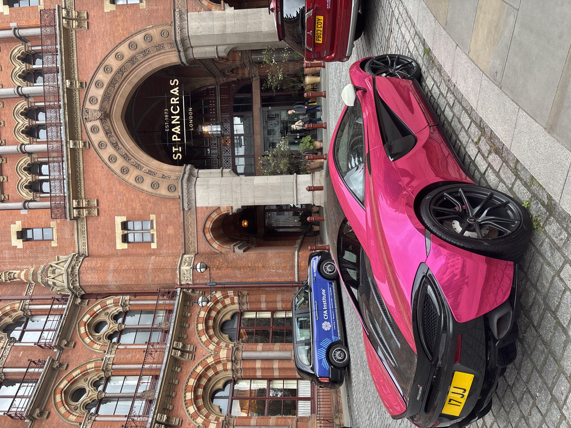 The image shows St. Pancras Renaissance Hotel in London, United Kingdom, with two cars parked on the street in front of the building. One car is a pink McLaren, and the other is a blue CFA Institute vehicle. A red vehicle is also partially visible behind the pink McLaren. People are standing and walking near the entrance of the hotel. The street is paved with grey bricks. The hotel’s facade features arched doorways and windows, and the name "St. Pancras" is prominently displayed above the entrance.