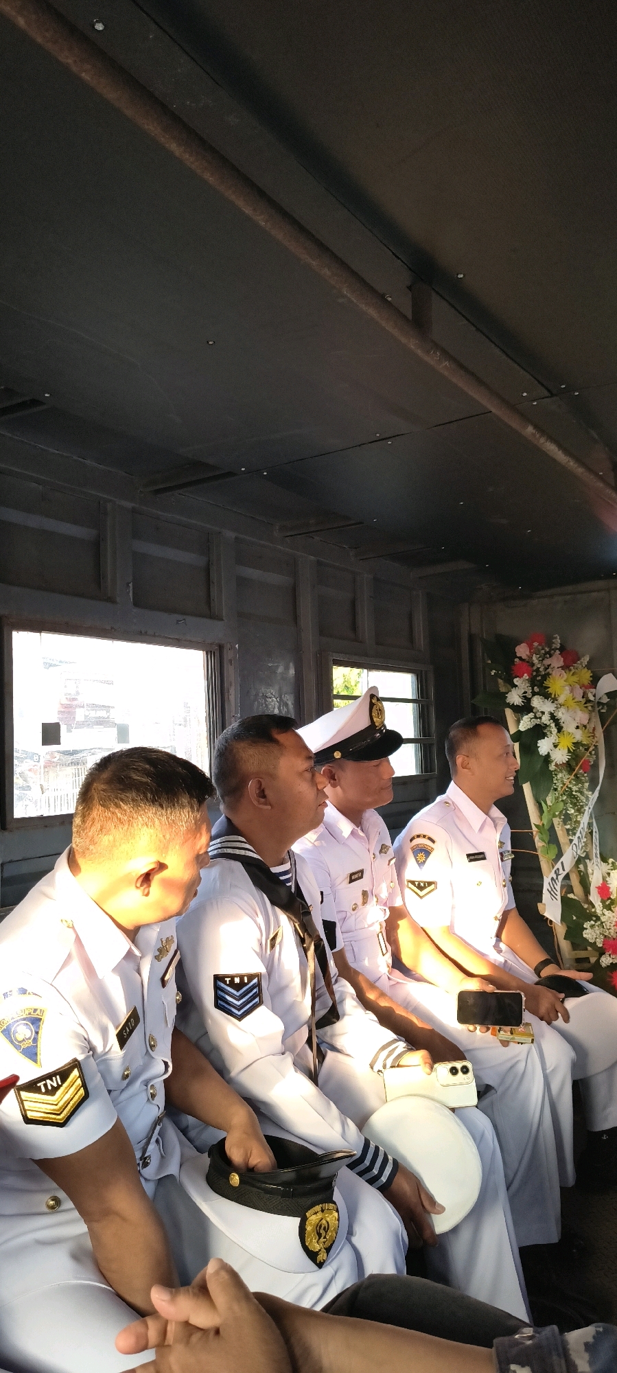 Four Indonesian Navy officers in white uniforms are seated in a vehicle. The officers are positioned in profile, facing right. The officer on the far left has a name tag that reads "SAID". The officer next to him wears a dark neckerchief. The officer in the center wears a white peaked cap. The officer on the far right is smiling and holding a smartphone. In the background, a floral arrangement is visible, featuring red, yellow, and white flowers. The vehicle's interior appears to be made of dark wooden panels and has two windows. A rusty pipe runs along the ceiling of the vehicle. The lighting suggests it is daytime, with sunlight casting shadows. The scene is indoors, within a vehicle, possibly in the city of Cirebon, Indonesia, judging by the uniforms and context of Indonesian military personnel.
