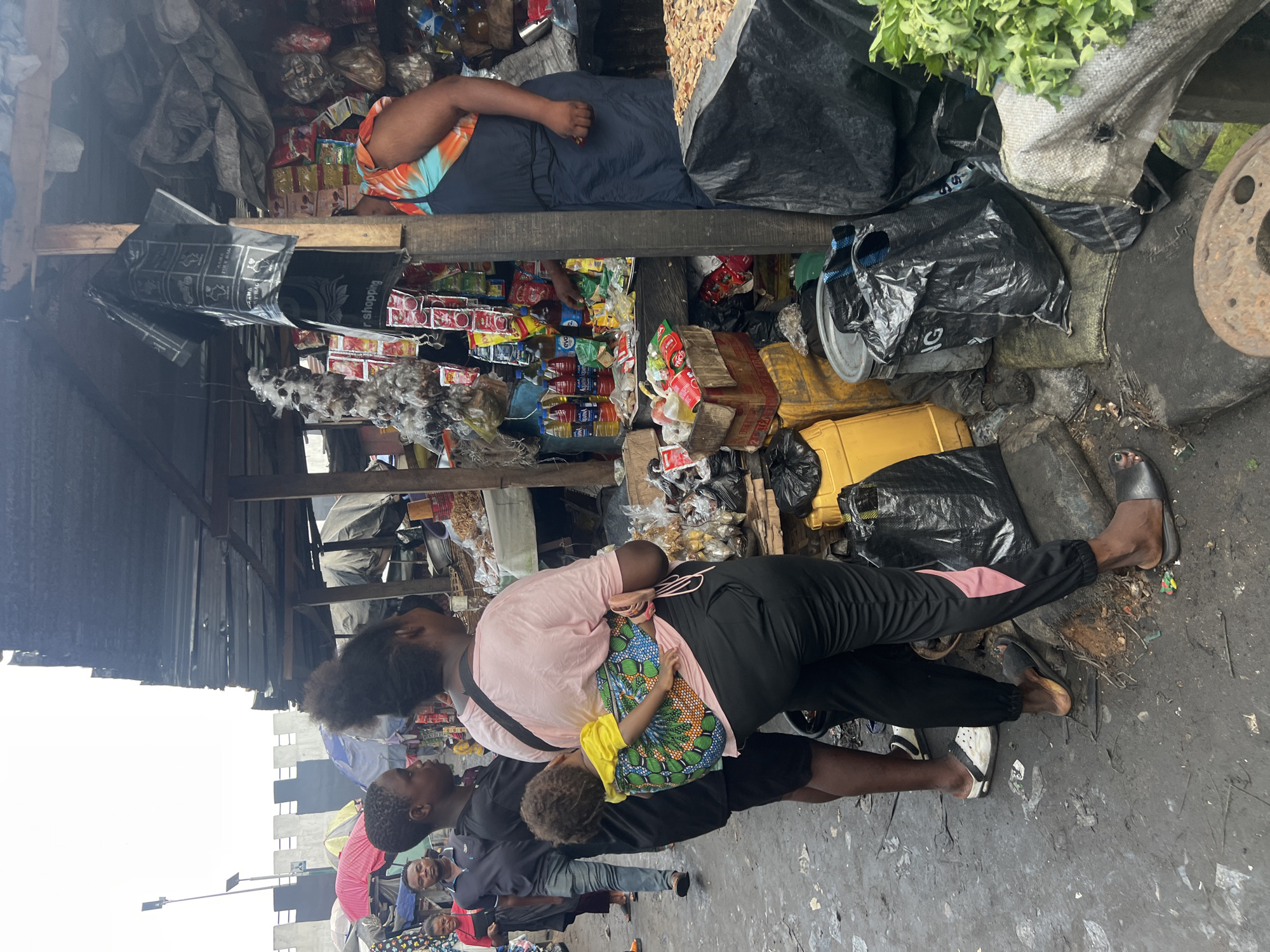 The image depicts a busy market scene in Nkpor, Nigeria, likely during daytime given the natural lighting. A group of people are visible, with a woman carrying a young child on her back. The child is wrapped in a colorful cloth, and the woman is wearing a pink t-shirt and black pants. Another person, likely a man, is standing next to them, wearing a dark shirt and gray pants. Other individuals are seen in the background, some appearing to be vendors or shoppers.
The setting is a market stall with various goods displayed, including what appears to be packaged food items, drinks, and fresh produce. Bags and other market materials are also visible, creating a somewhat cluttered but vibrant atmosphere. The ground is dusty and appears unpaved.
One of the black bags has some text in white, though it is partially obscured and difficult to read clearly. The overall mood seems to be one of everyday activity and commerce. The image is taken from a slightly elevated angle, looking down on the scene.