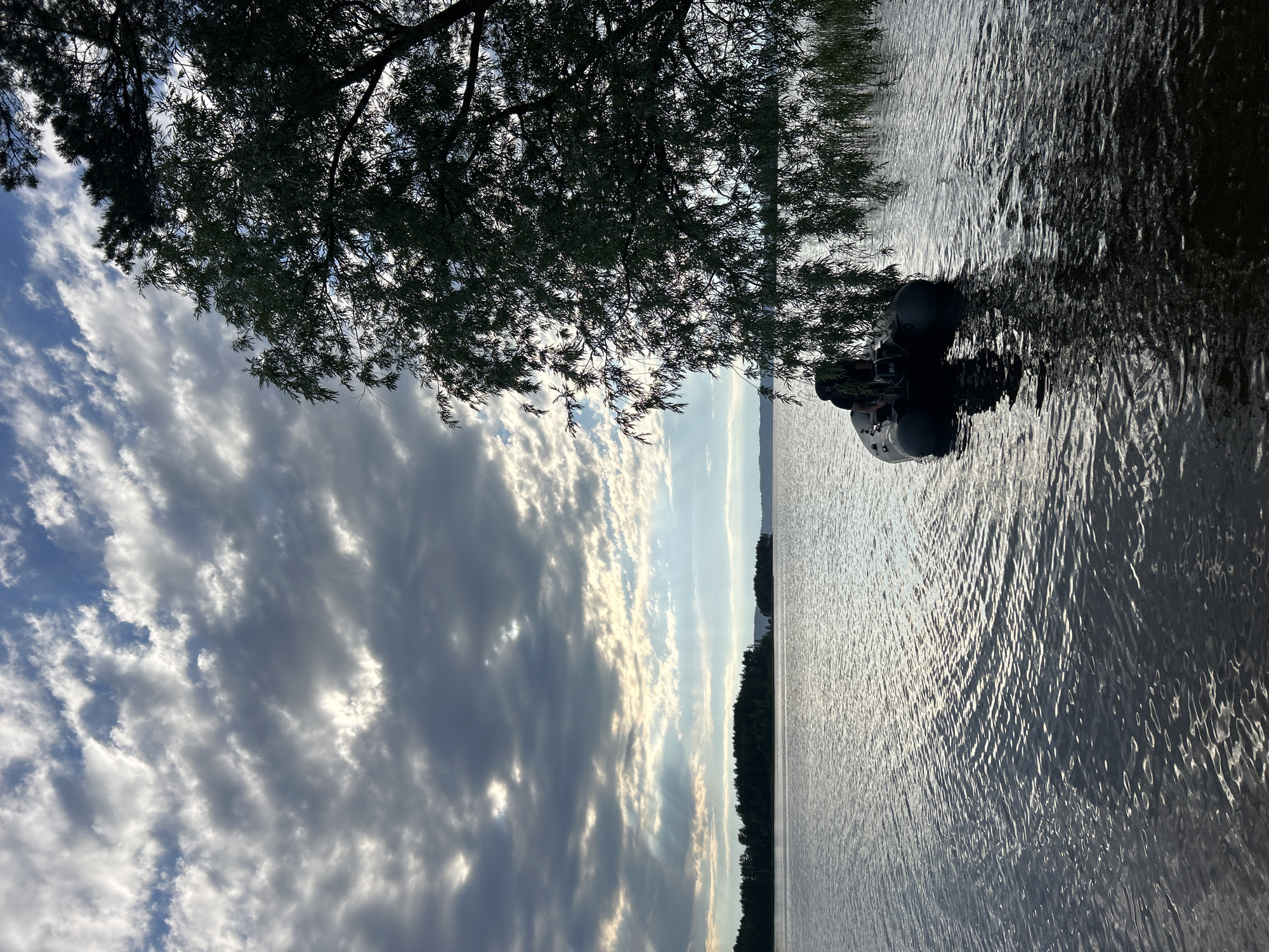 The image depicts a tranquil scene on a body of water, likely a lake, near Vekoshinka, Russia. In the foreground, a dark-colored inflatable boat with an outboard motor is moored on the water. A person, dressed in dark clothing and wearing a backpack, is seated in the boat. The boat is positioned near the shore, where the branches of a large tree, with drooping leaves, hang over the water. The water surface is calm with gentle ripples reflecting the light. In the background, a distant shoreline is visible, covered with a dense forest. The sky above is dramatic, with a mix of dark, heavy clouds and patches of bright blue sky, through which rays of sunlight break through, illuminating parts of the water and the distant landscape. The overall atmosphere is one of natural beauty and solitude.
