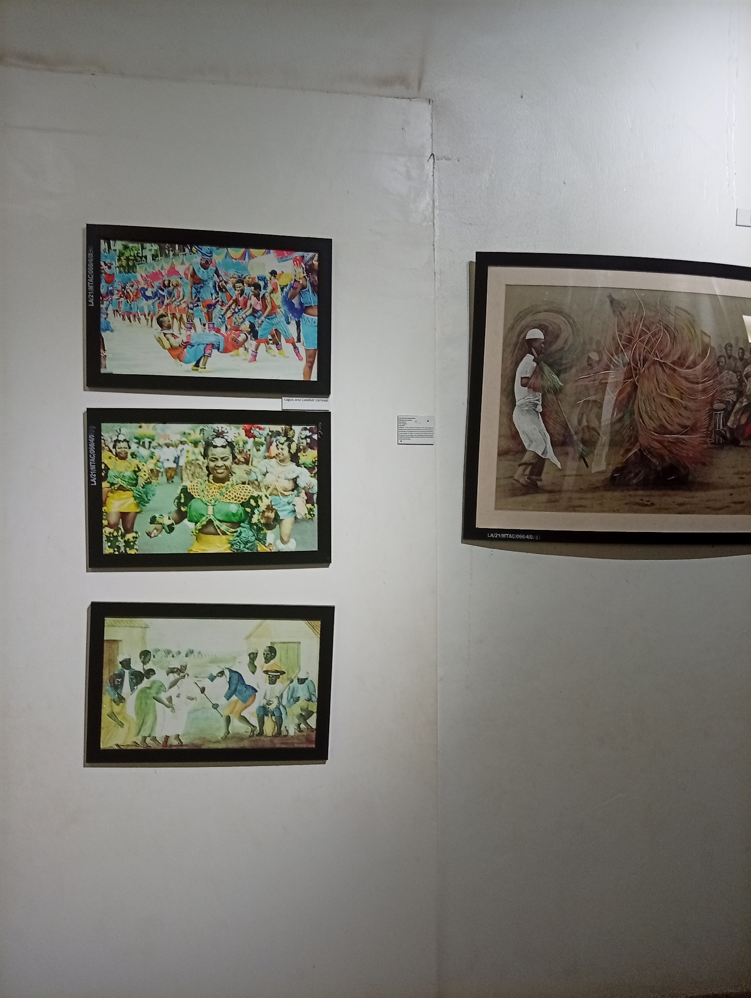 The image depicts a display of framed pictures on a white wall, possibly within an art gallery or cultural center in Badagry, Nigeria. On the left wall, there are three rectangular framed pictures stacked vertically. The top picture shows a lively carnival scene with people in colorful costumes. The middle picture features individuals in traditional attire, one of whom wears a green ensemble and a large neck adornment. The bottom picture depicts a scene with people dressed in older attire, possibly depicting a historical event or cultural tradition.
On the right wall, another framed picture is partially visible. It appears to be an illustration of a man in white clothing holding an object next to another individual in elaborate brown attire. A small informational card is affixed to the wall next to this picture. The corner where the two walls meet creates a visible line. The lighting is diffused, casting a soft glow on the scene.